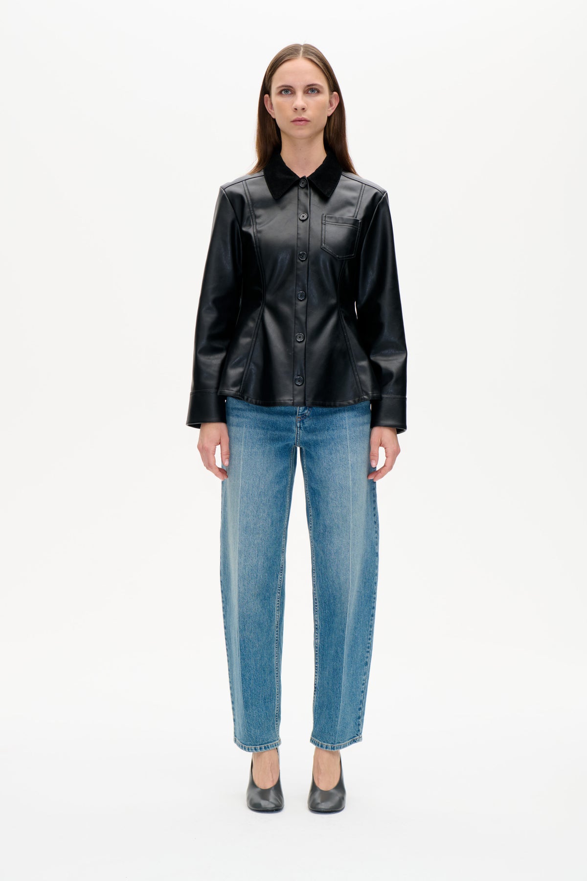 Front view of a model wearing a black button-front jacket with a contrasting collar and chest pocket, blue jeans and black shoes.
