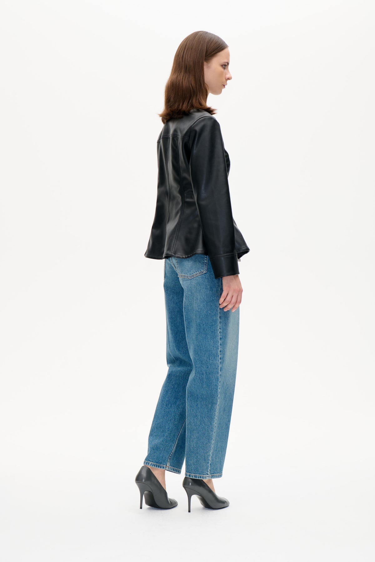 Side-profile model wearing a glossy black jacket with a brown textured collar, blue jeans and black heels.