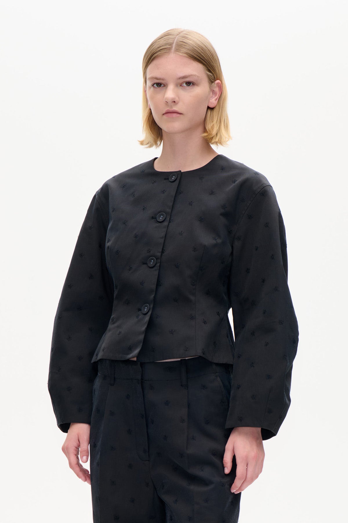 Model iført en sort croppedjakke med diskrete broderede motiver, rund hals og store knapper, kombineret med matchende bukser.