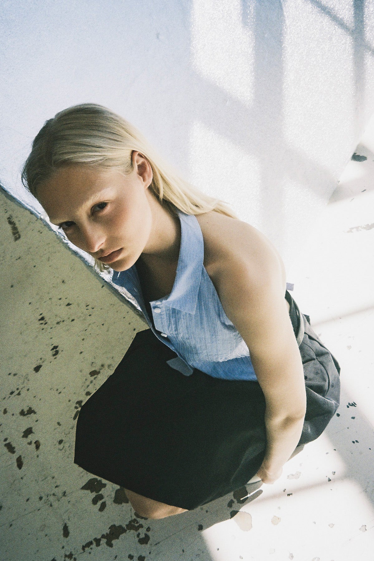 En model læner sig frem iført en lyseblå ærmeløs top med krave og knapper foran og en sort nederdel, fotograferet oppefra.