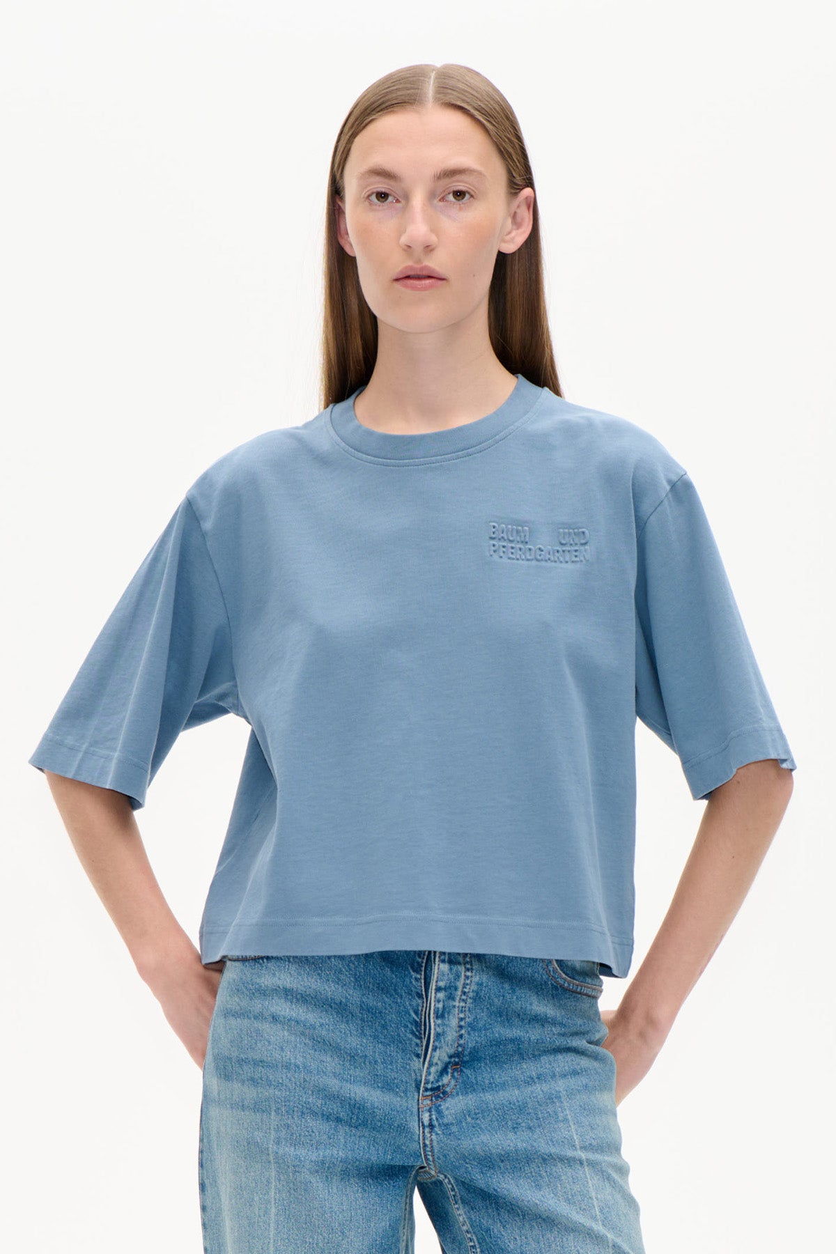 Forfra af en model iført en lyseblå oversized kortærmet t-shirt med et præget logo på brystet og blå jeans.