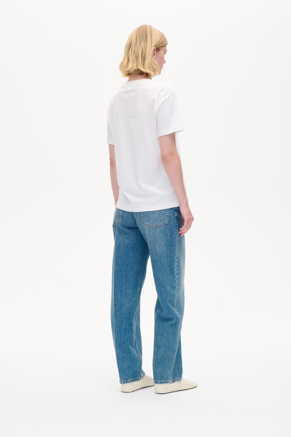 Set bagfra: en model iført en ensfarvet hvid t-shirt med korte ærmer, blå jeans og hvide sko mod en hvid baggrund.