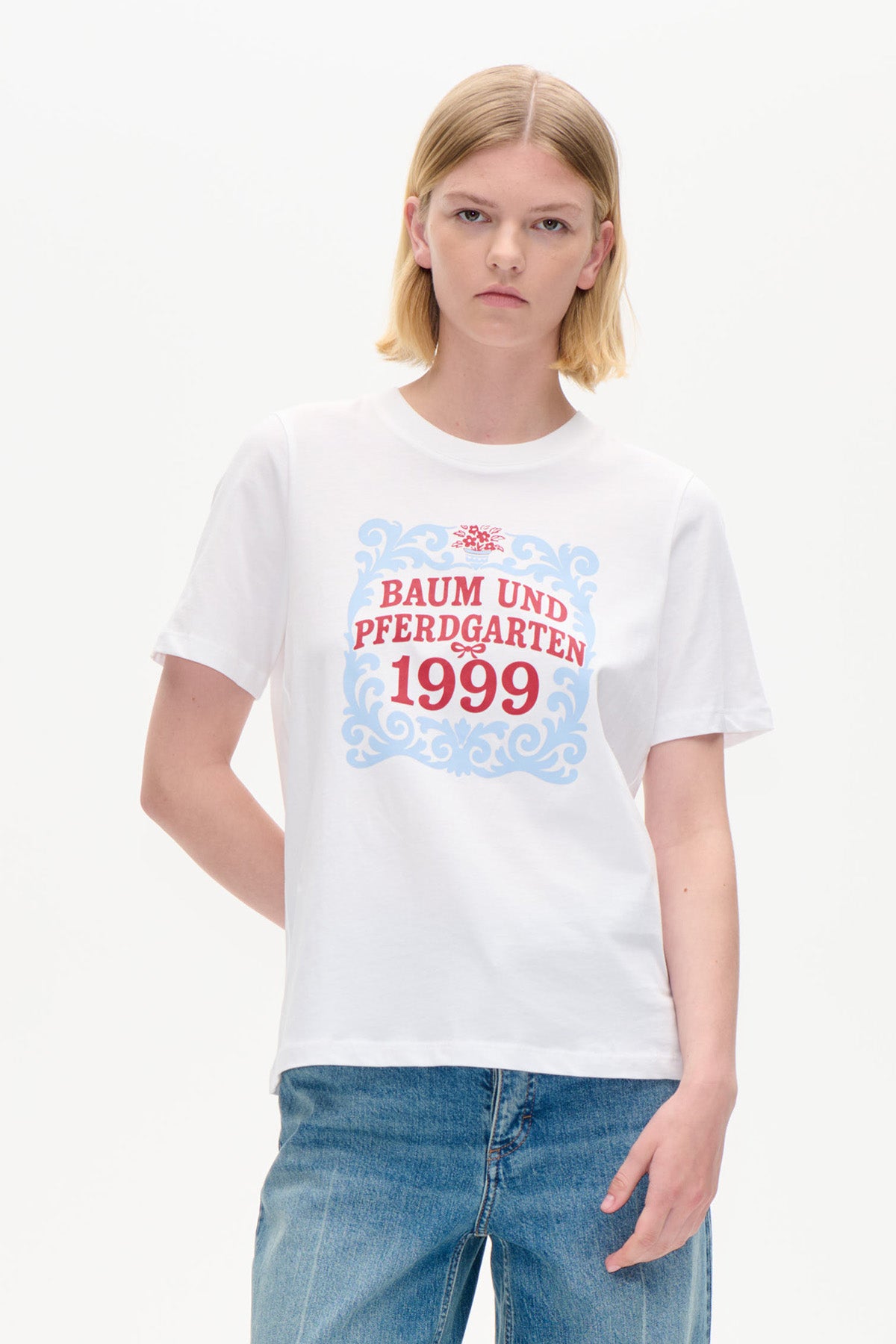 Hvid kortærmet t-shirt med en blå ornamentramme og en rød Baum und Pferdgarten 1999-grafik, set forfra.