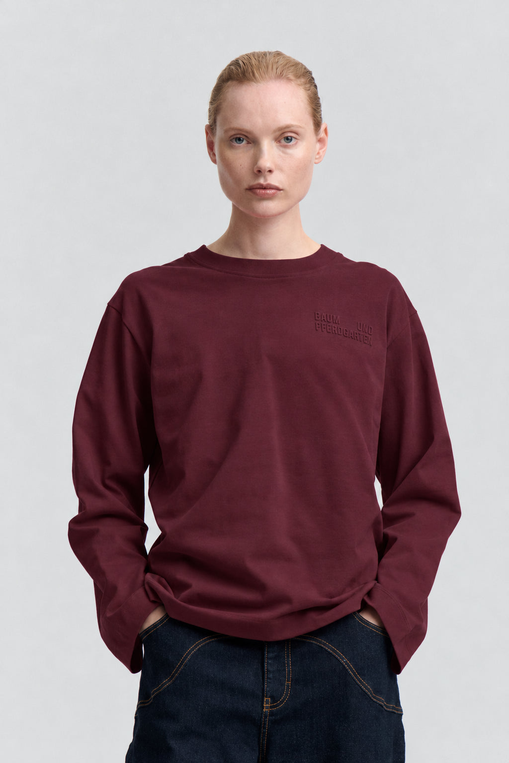 Set forfra af en bordeaux langærmet sweatshirt med rund hals og diskrete ton-i-ton bogstaver på brystet, stylet med mørke jeans.