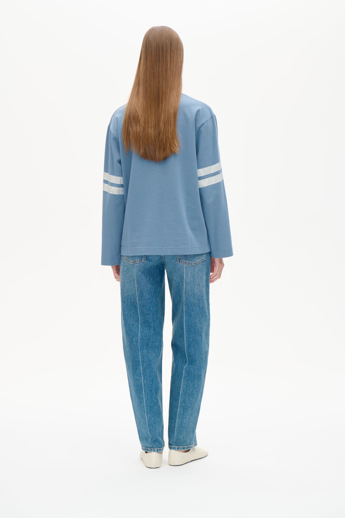 Set bagfra: en model iført en lyseblå langærmet sweatshirt med hvide striber på ærmerne og blå jeans.