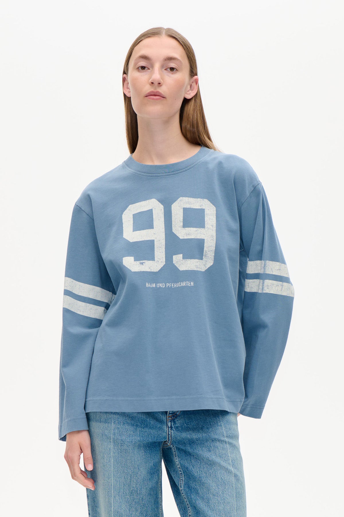 Model iført en støvetblå langærmet sweatshirt med hvidt "99" brystprint og dobbelte ærmestriber, stylet med jeans.