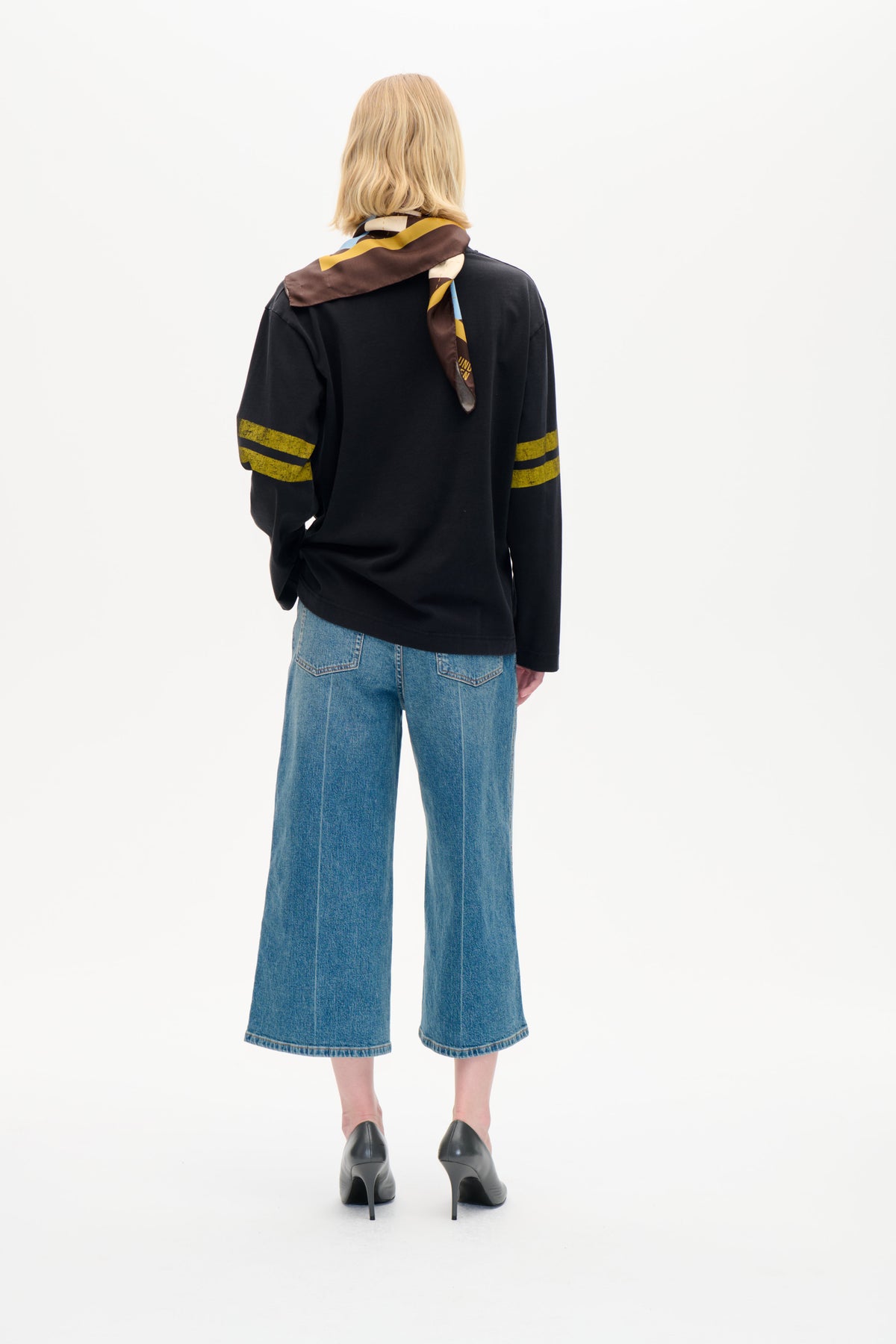 Set bagfra af en model i en sort langærmet top med sennepsfarvede striber på ærmerne, et flerfarvet geometrisk tørklæde og blå wide-leg jeans.