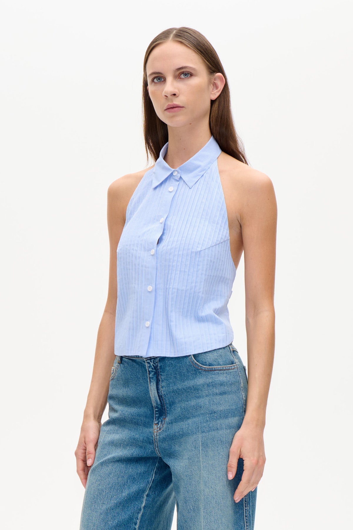Model iført en lyseblå, fintstribet ærmeløs top med krave, knapper foran, halterneckudskæring og blå jeans.