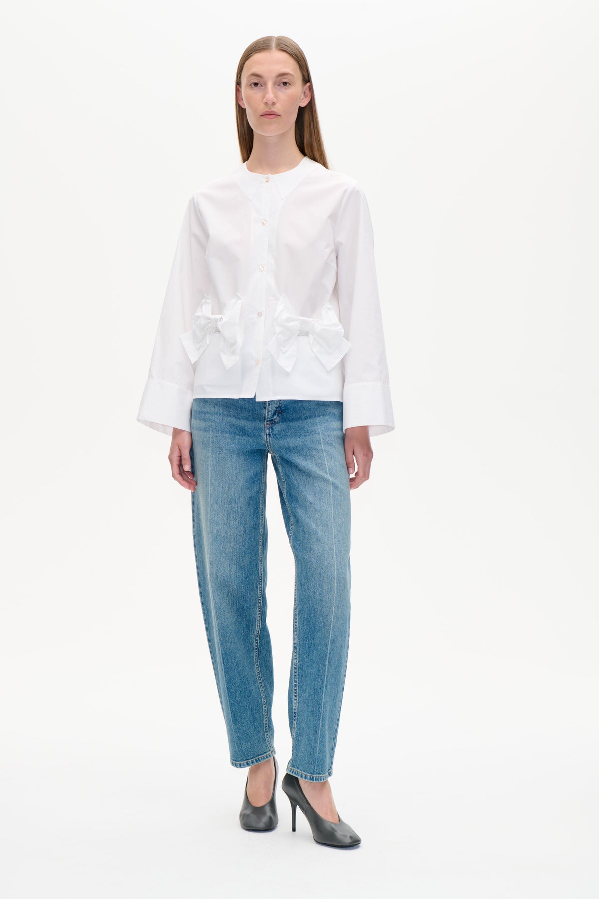 Set forfra: en hvid skjorte med knapper foran, oversized spids krave og skulpturelle sløjfedetaljer i taljen, kombineret med blå jeans.