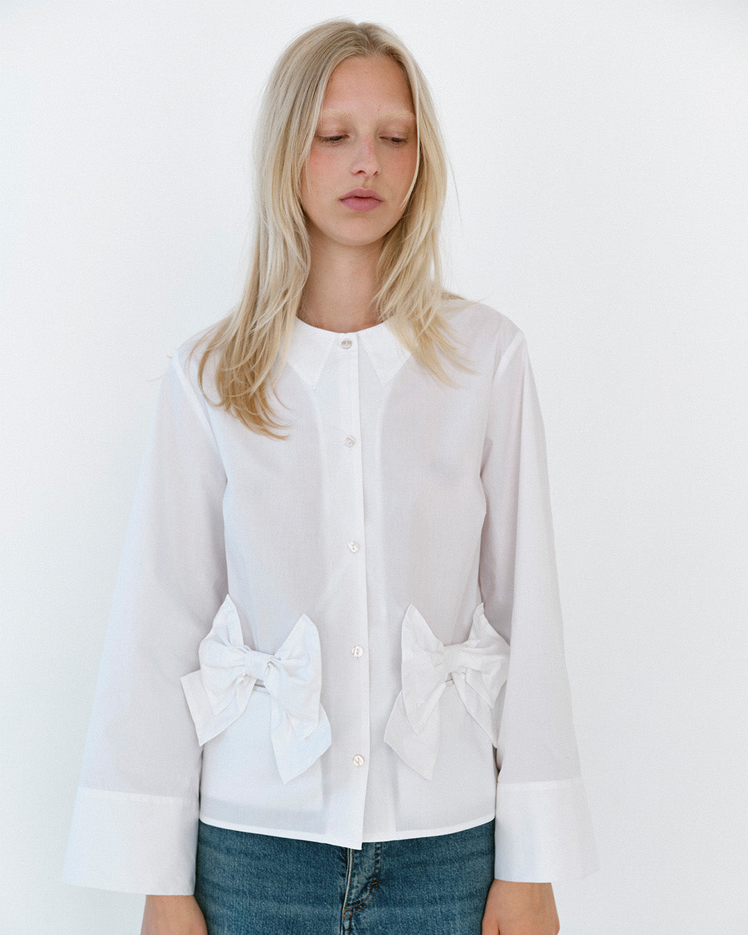 Model iført en hvid bluse med knapstolpe, spids krave, oversized sløjfedetaljer i taljen og vide manchetter, stylet med blå jeans.