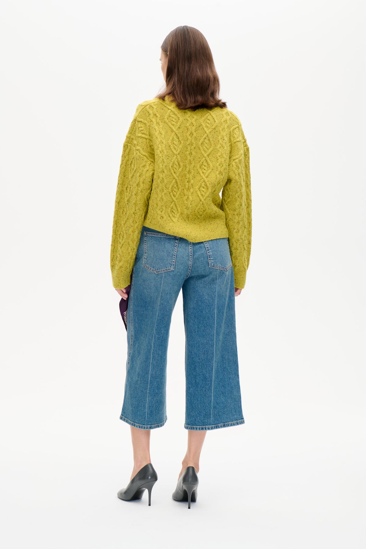 Bagfra ses en model iført en gulgrøn, meleret kabelstrikket sweater og blå cropped, vidbenede jeans.