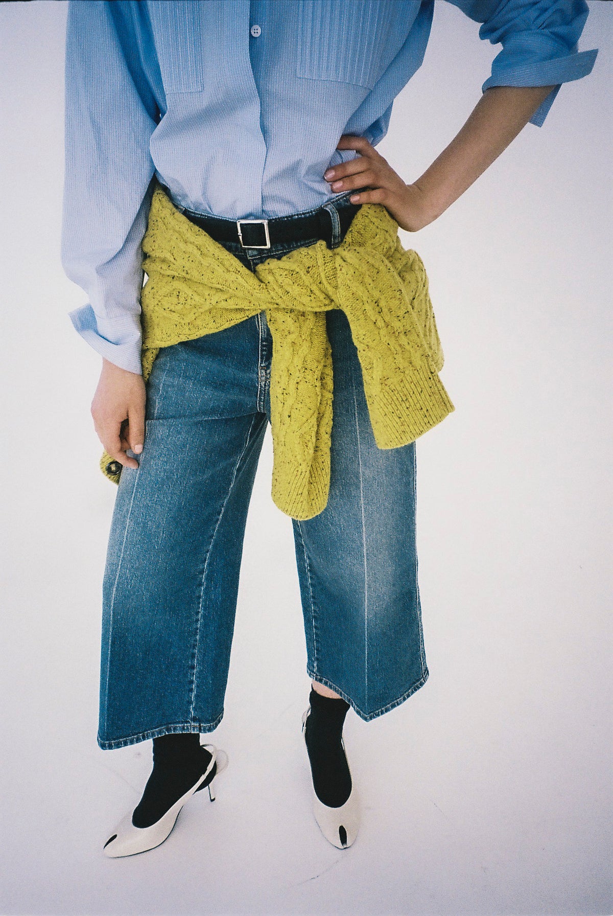 Cropped kampagnebillede af en lyseblå button-up skjorte, blå jeans med vide ben og en sennepsgul Sweater bundet i taljen.