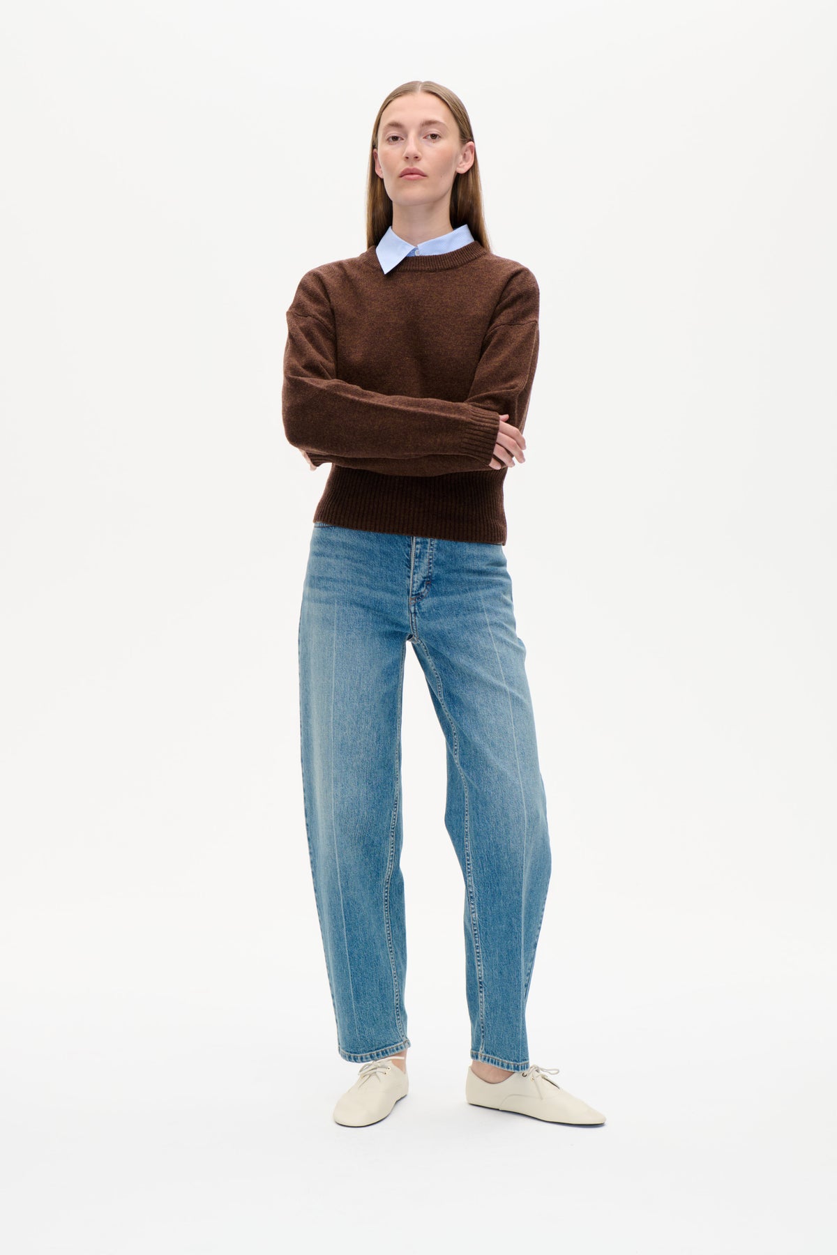 Forfra af en model iført en brun rundhalset Sweater over en lyseblå, stribet skjorte med krave, blå jeans og cremefarvede snøresko.