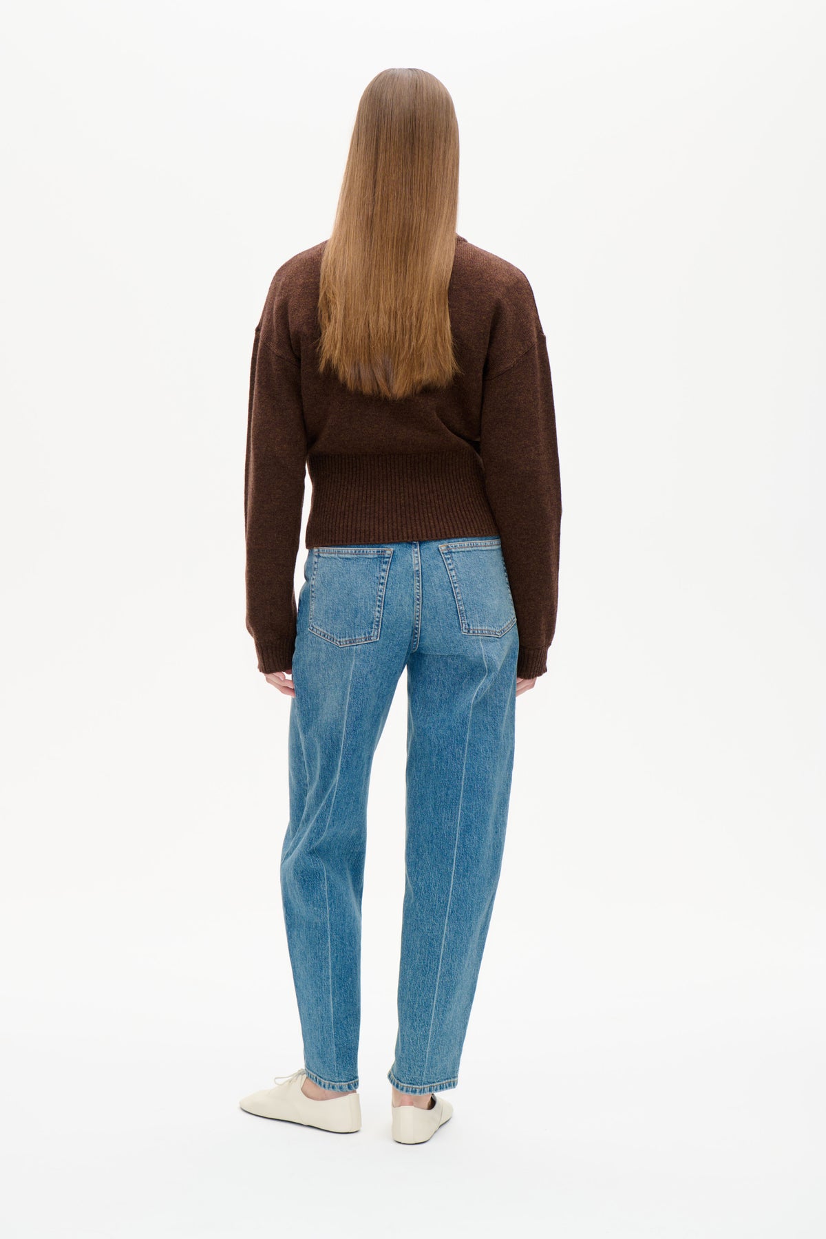 Set bagfra af en model iført en mørkebrun langærmet strikket Sweater med ribkant og blå jeans.