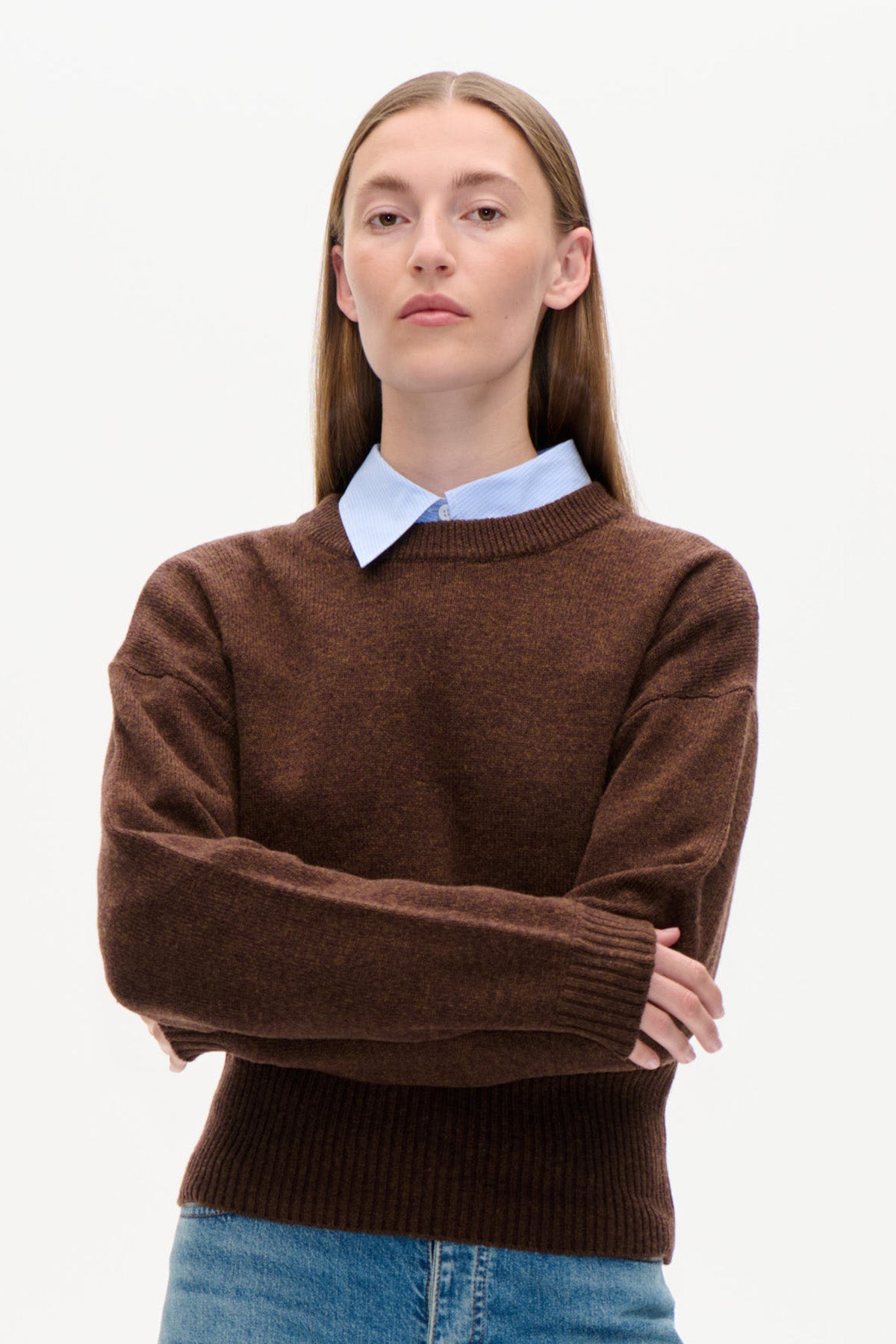 Forfra billede af en model iført en brun rundhalset strikket sweater over en lyseblå skjorte med krave og blå jeans.