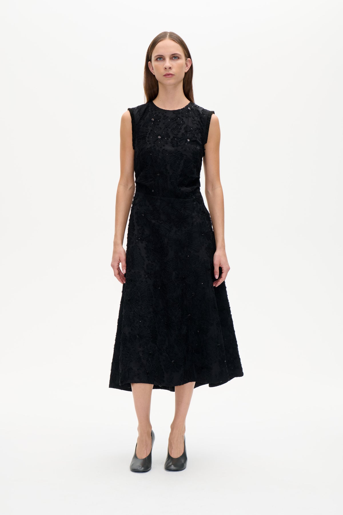 Model set forfra iført en sort ærmeløs kjole med ophøjede blomsterdetaljer og spredte palietter, indsnævret talje og skørt med vidde.