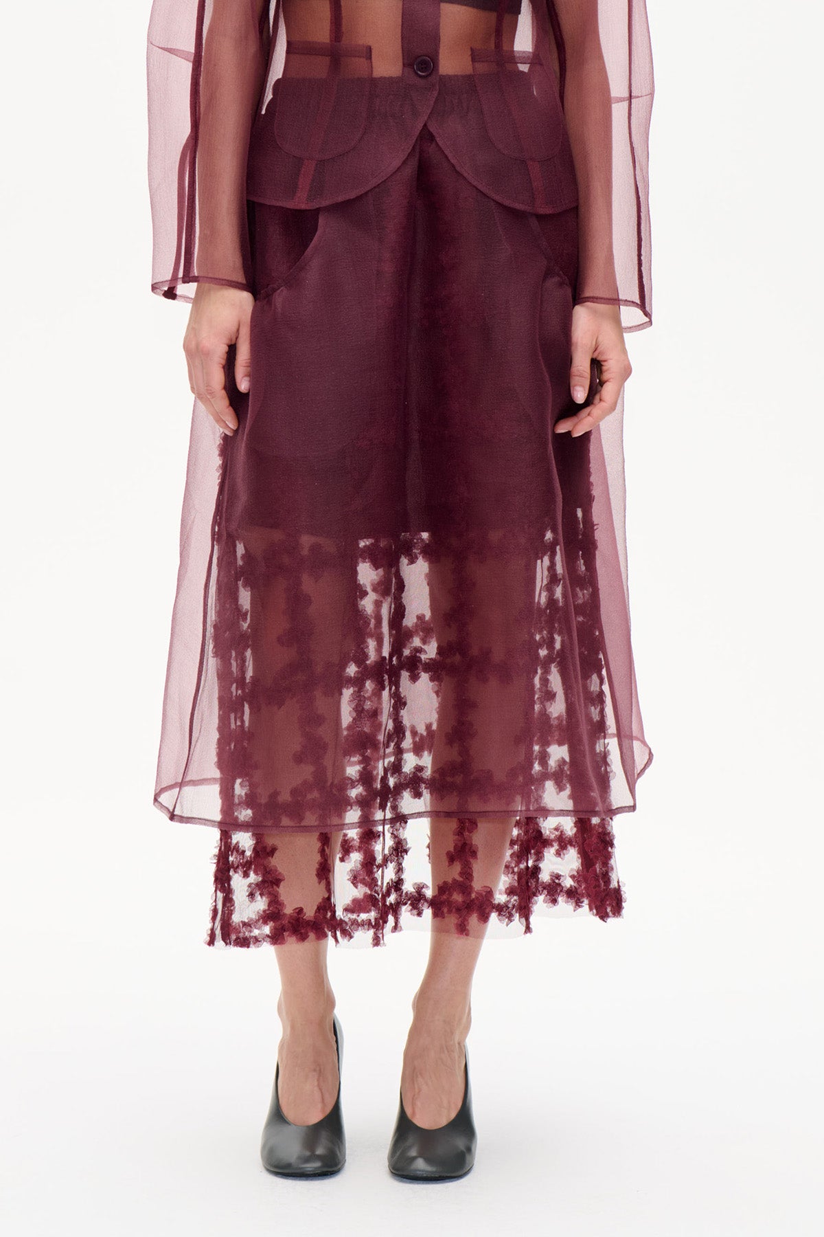 Cropped front view of a model wearing a deep burgundy skirt with a sheer floral-patterned hem and a matching translucent jacket with rounded patch pockets.