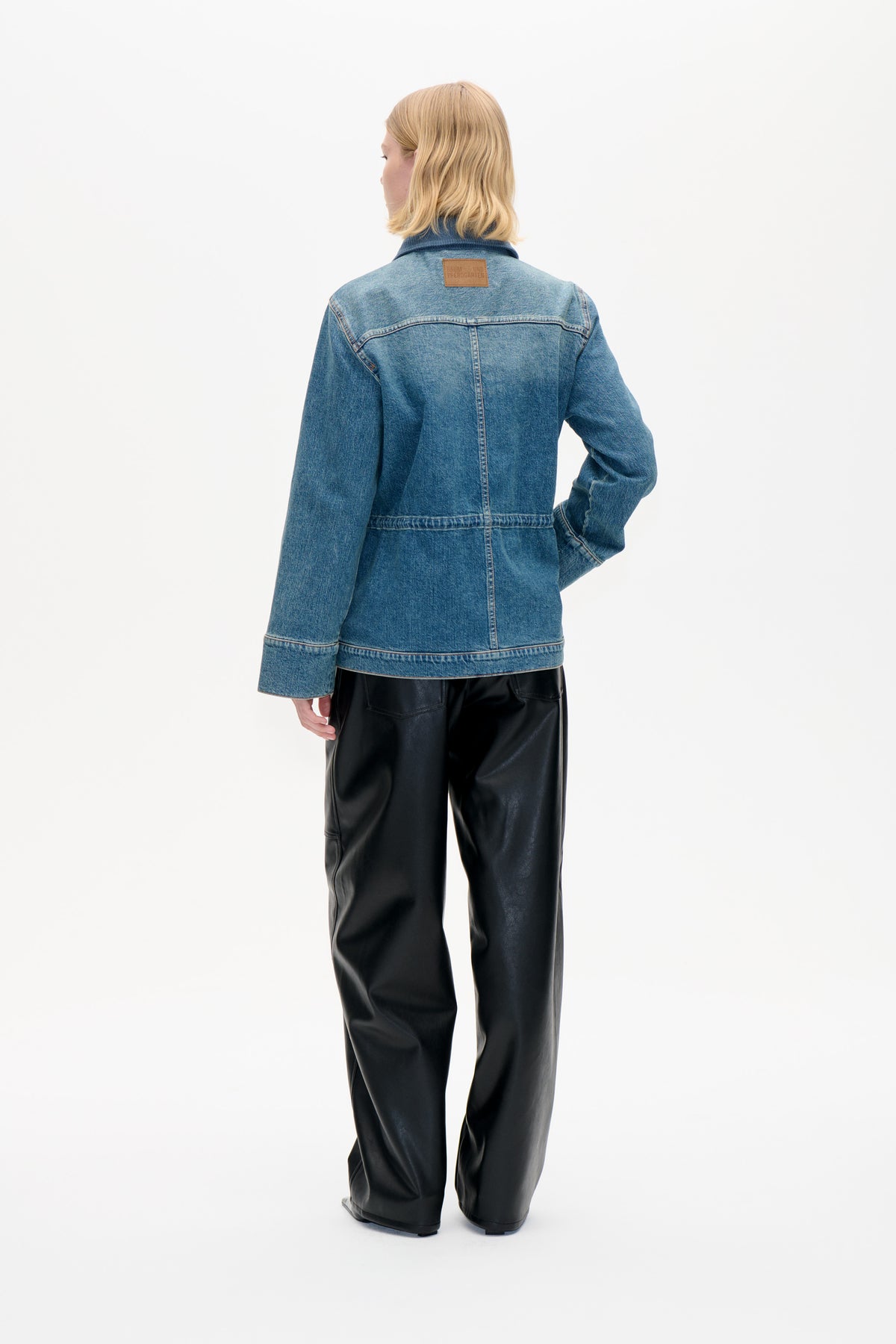Set bagfra: en model iført en blå denimjakke med mørk ribbet krave og lysebrun mærkepatch, kombineret med sorte bukser.