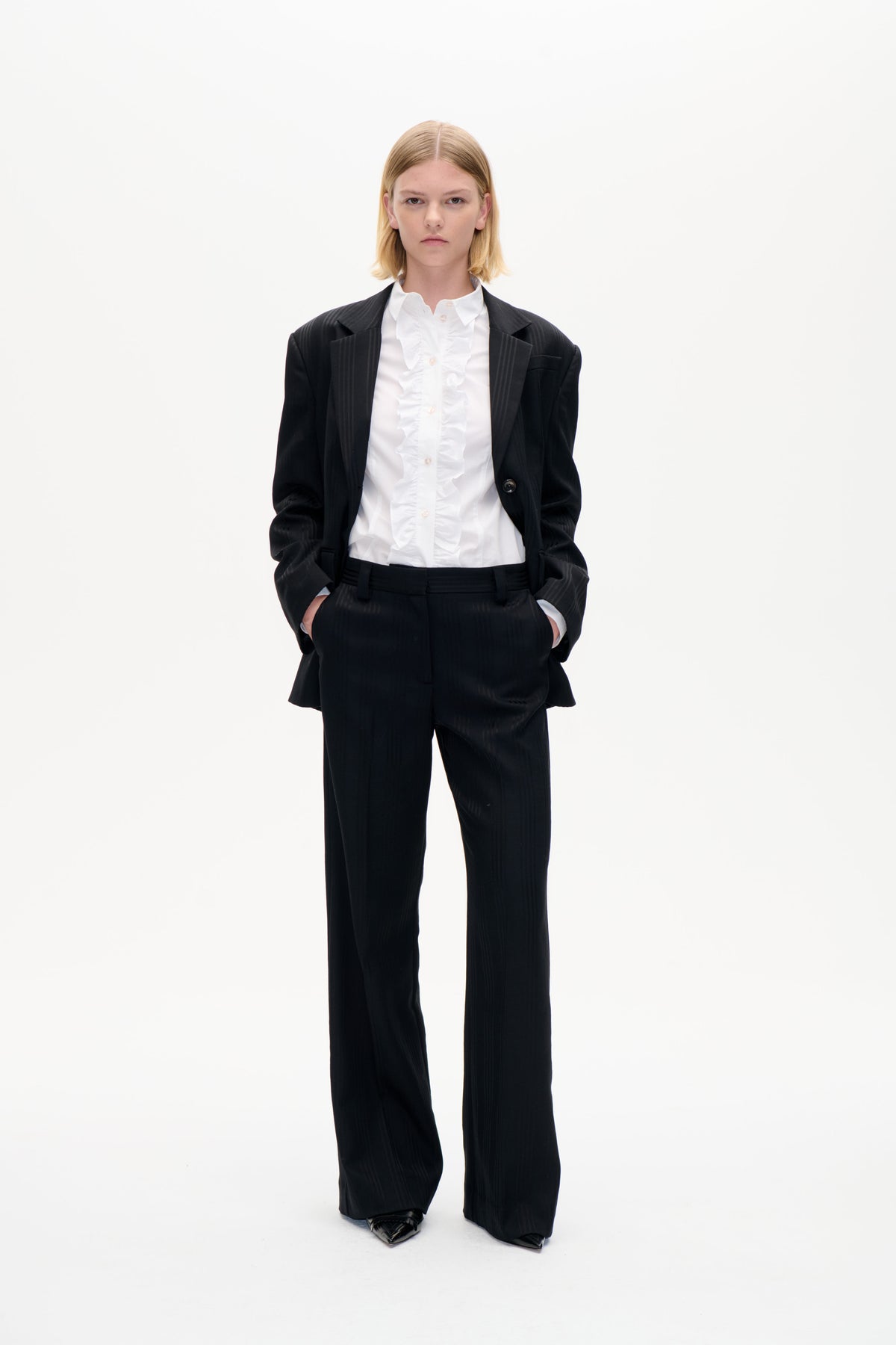 Model set forfra i en sort nålstribet blazer og bukser med vide ben over en hvid skjorte med flæser, mod en ensartet hvid baggrund.