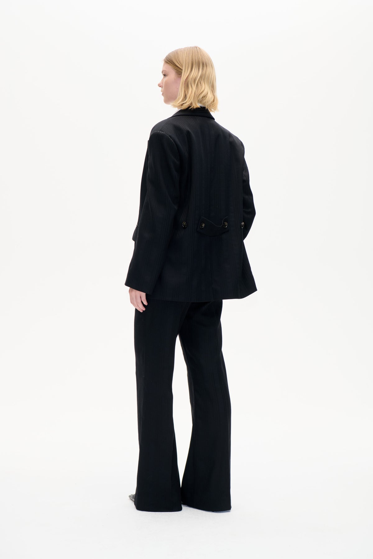 Model i en sort blazer med tynde kridhvide striber, en knappet bagstrop i taljen og bukser med vide ben, vist fra siden og bagfra.