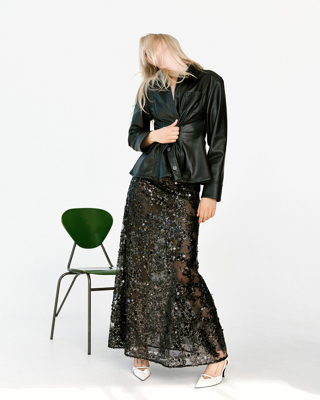 Model in a glossy black button-front jacket and sheer black sequin maxi skirt, standing beside a green chair in white heels.