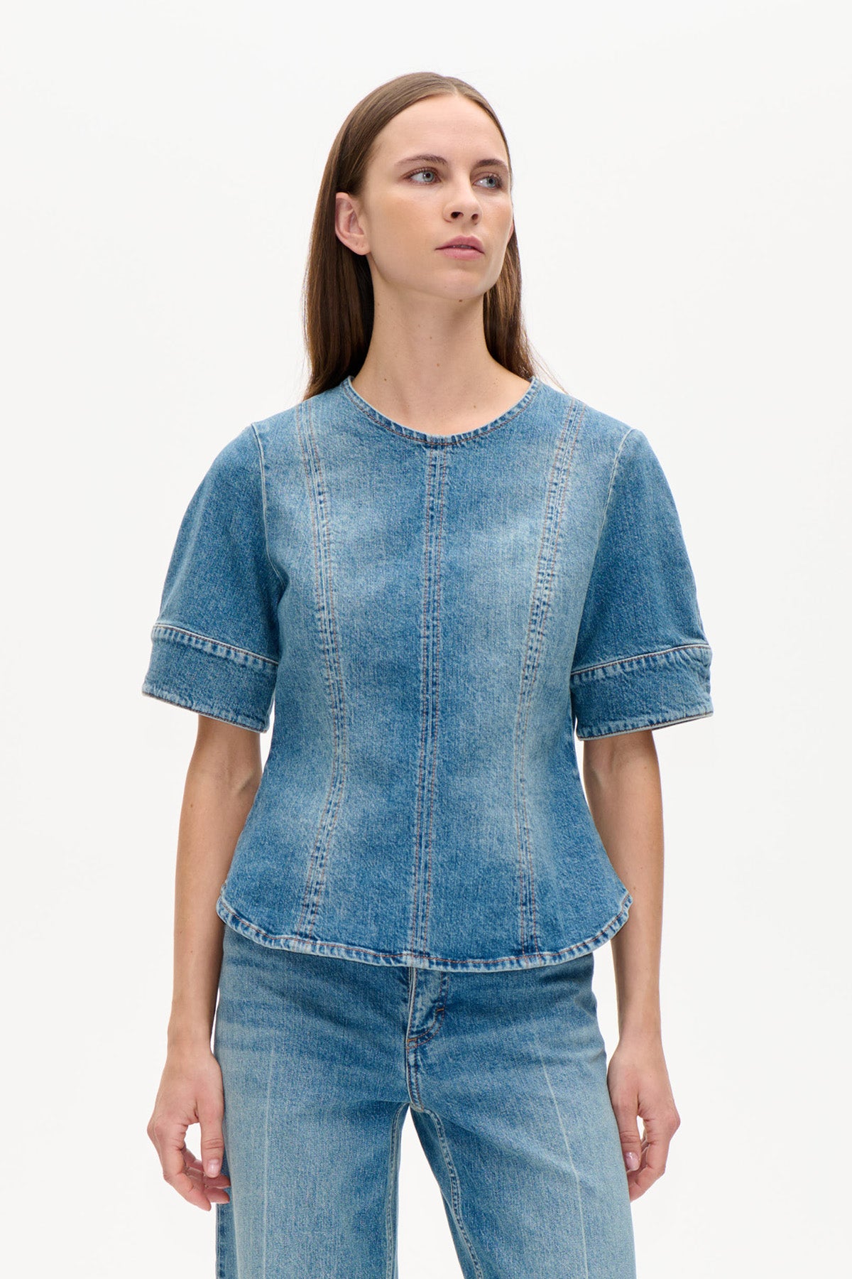 Front view of a blue denim top with a round neckline, short wide sleeves, and vertical panel stitching, worn with jeans.