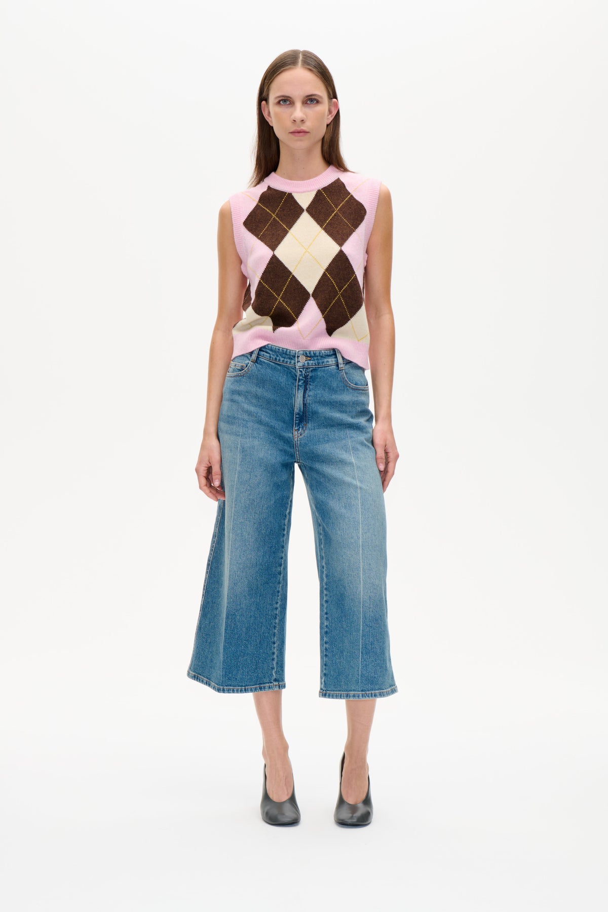 Forfra: en model iført en ærmeløs argyle-strikvest i pink, creme og brun samt blå wide-leg-jeans.