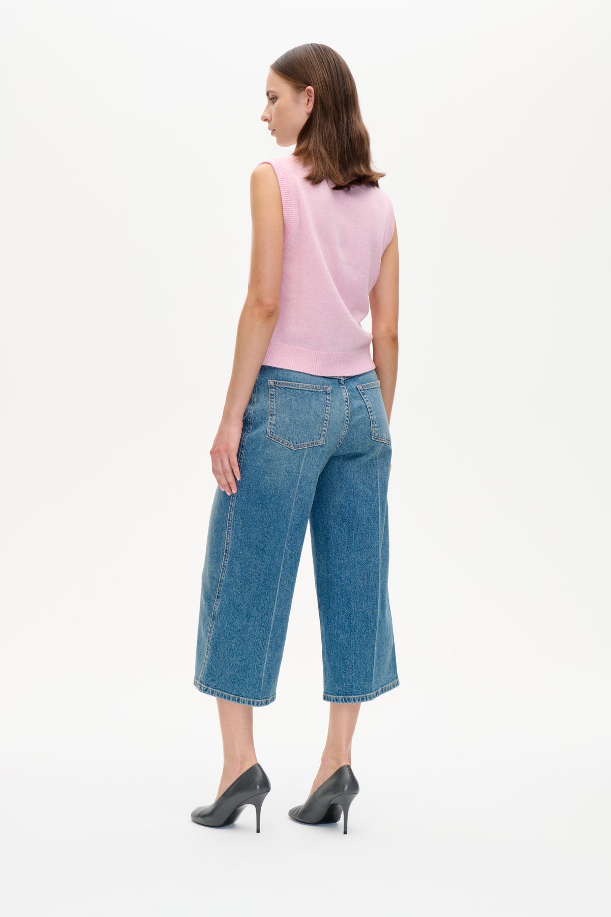 Model i en sart lyserosa ærmeløs strikvest og blå, vide croppedjeans, vist fra siden og bagfra.