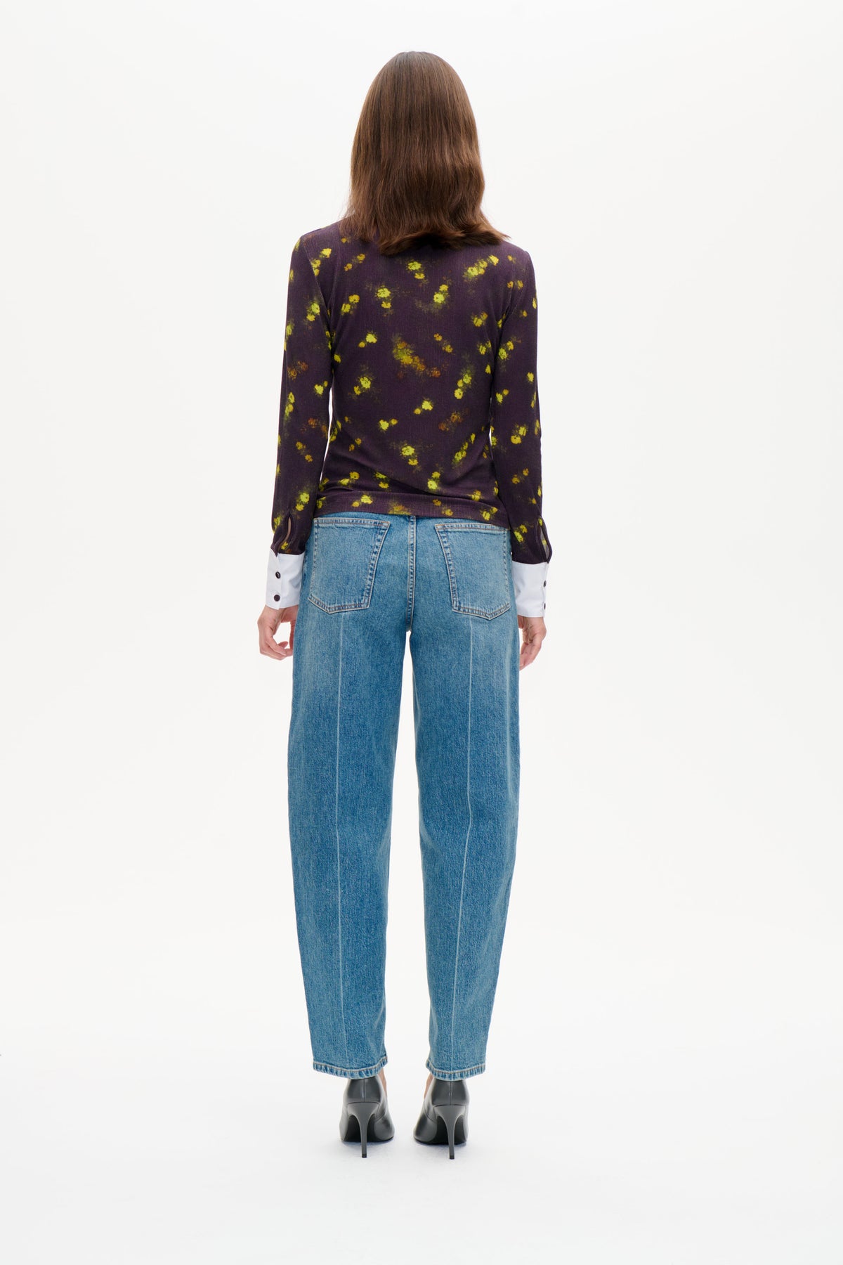 Back view of a model wearing a long-sleeved dark purple top with scattered yellow-green floral print, layered over white cuffs and blue jeans
