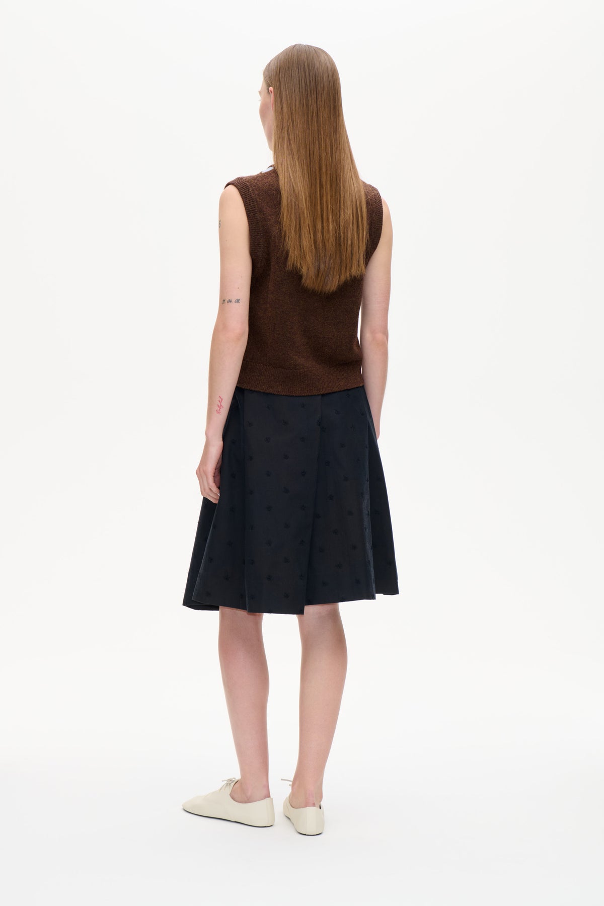 Set bagfra af en model iført en mørkebrun ærmeløs strikketop, en sort nederdel med diskrete motiver og cremefarvede ballerinaer.