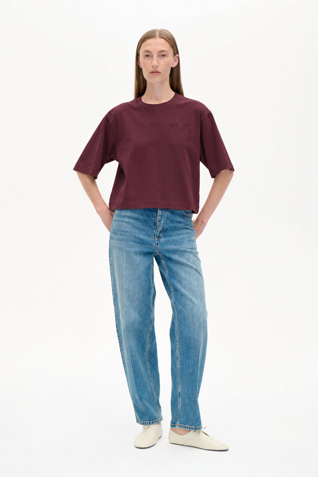 Forfra: en model iført en burgunder kortærmet t-shirt med tonet brystlogo og lyseblå mønstrede wide-leg-jeans.