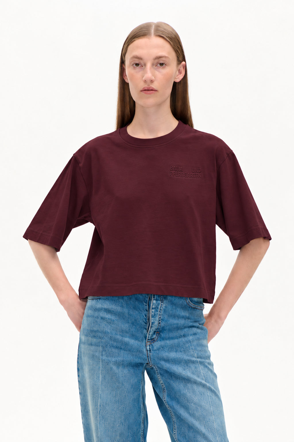 Model iført en cropped bordeaux kortærmet t-shirt med et præget logo på brystet og lyseblå mønstrede jeans.