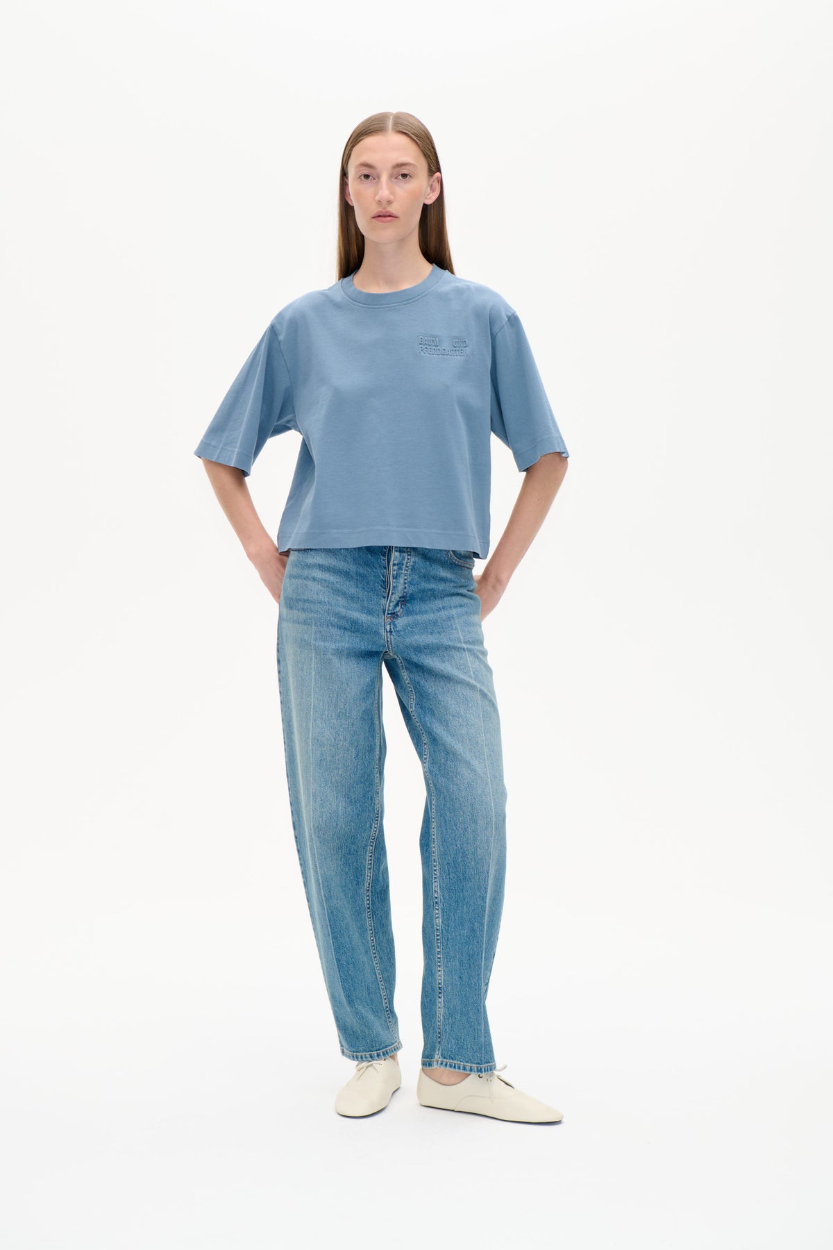 Model set forfra iført en oversized blå t-shirt med rund hals, albuelange ærmer og et ton-i-ton brystlogo, stylet med jeans.