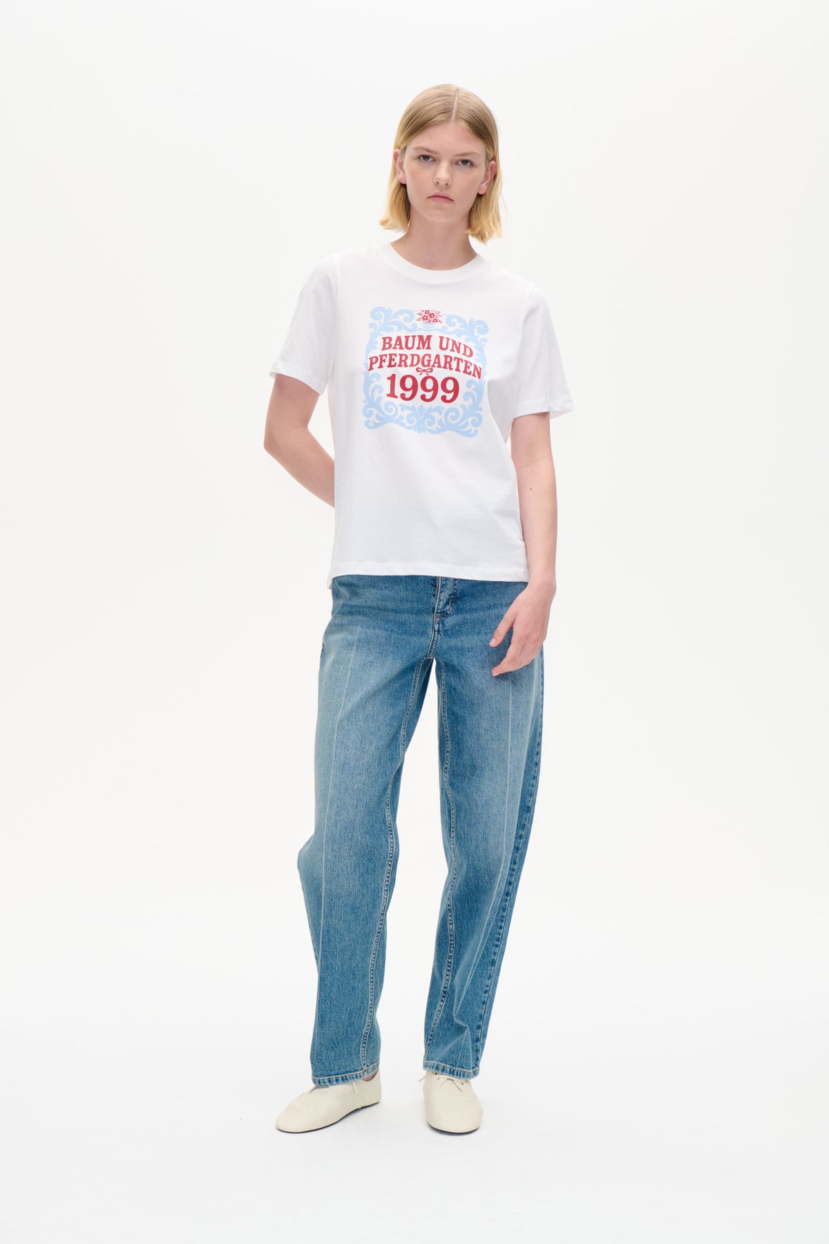 Model iført en hvid kortærmet t-shirt med et blå- og rødt ornamentalt print med teksten 'Baum und Pferdgarten 1999', stylet med blå jeans.