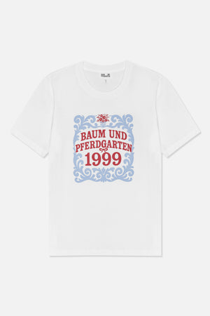 Forfra: en hvid kortærmet t-shirt med en blå ornamentramme, en rød blomsterdetalje og teksten 'Baum und Pferdgarten 1999'.