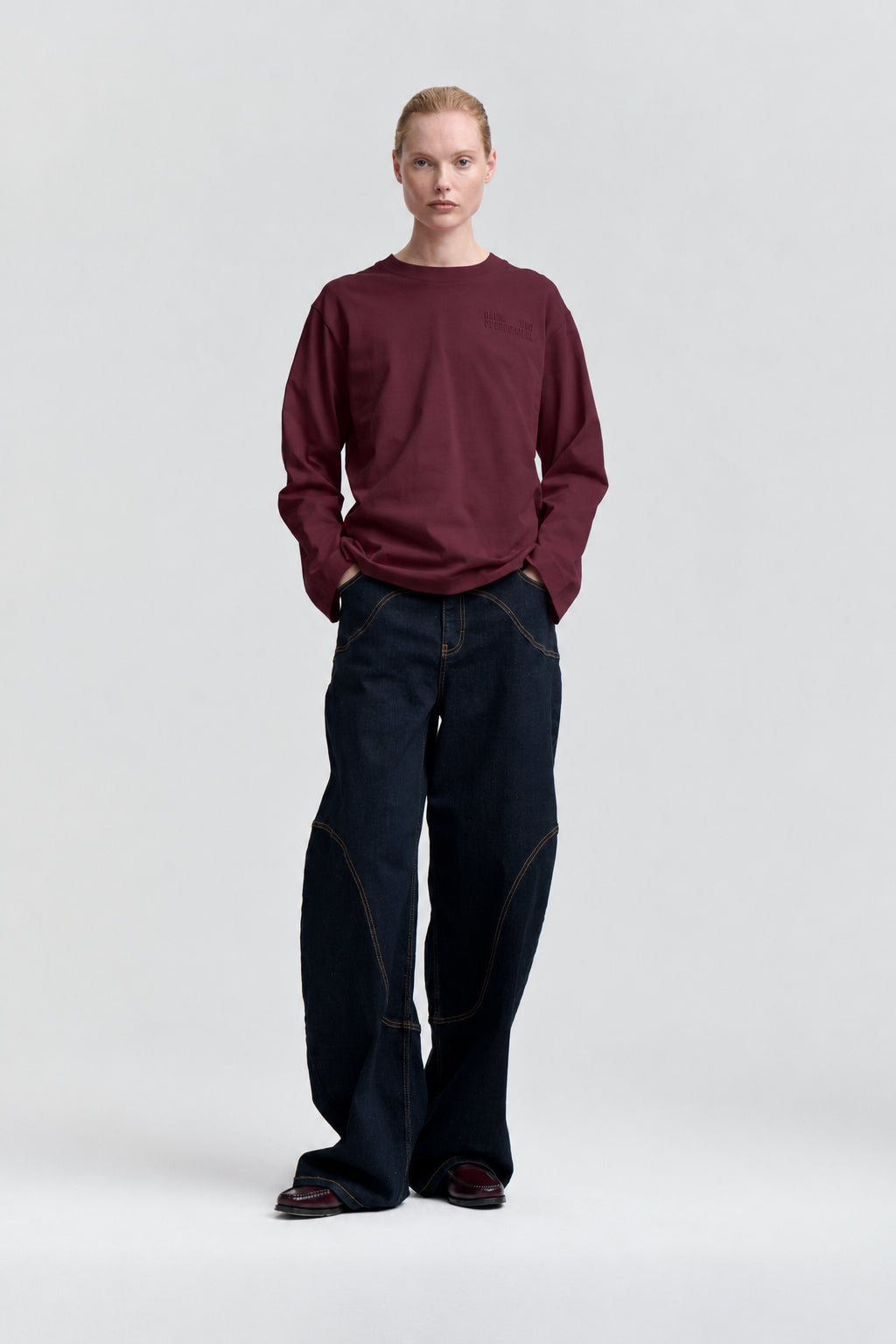 Model set forfra iført en bordeaux langærmet sweatshirt og vidbenede mørke indigojeans med kontrastsyning.