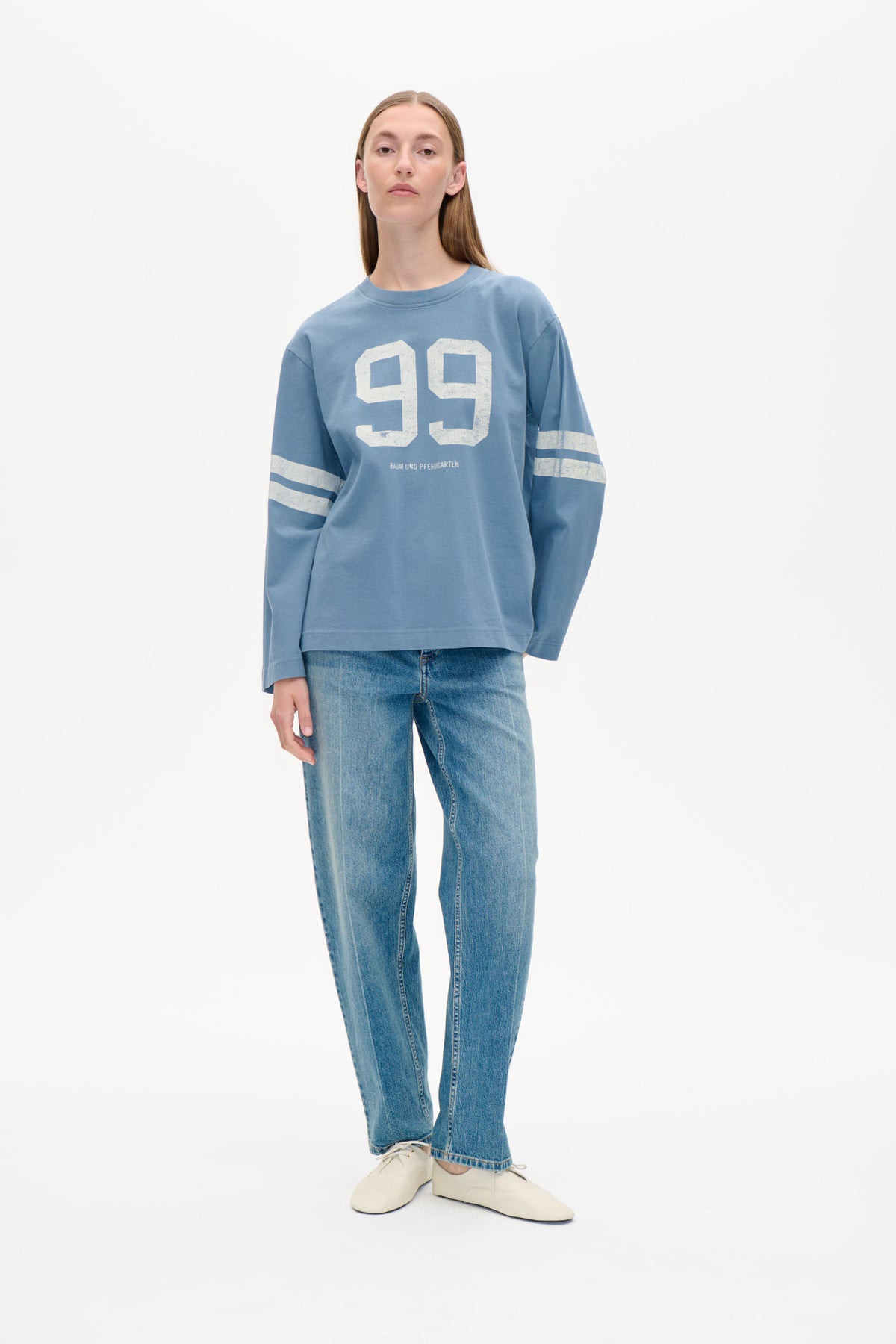 Forfra af en model iført en løs blå sweatshirt med en stor, slidt hvid 99-grafik og stribede ærmer, stylet med jeans.