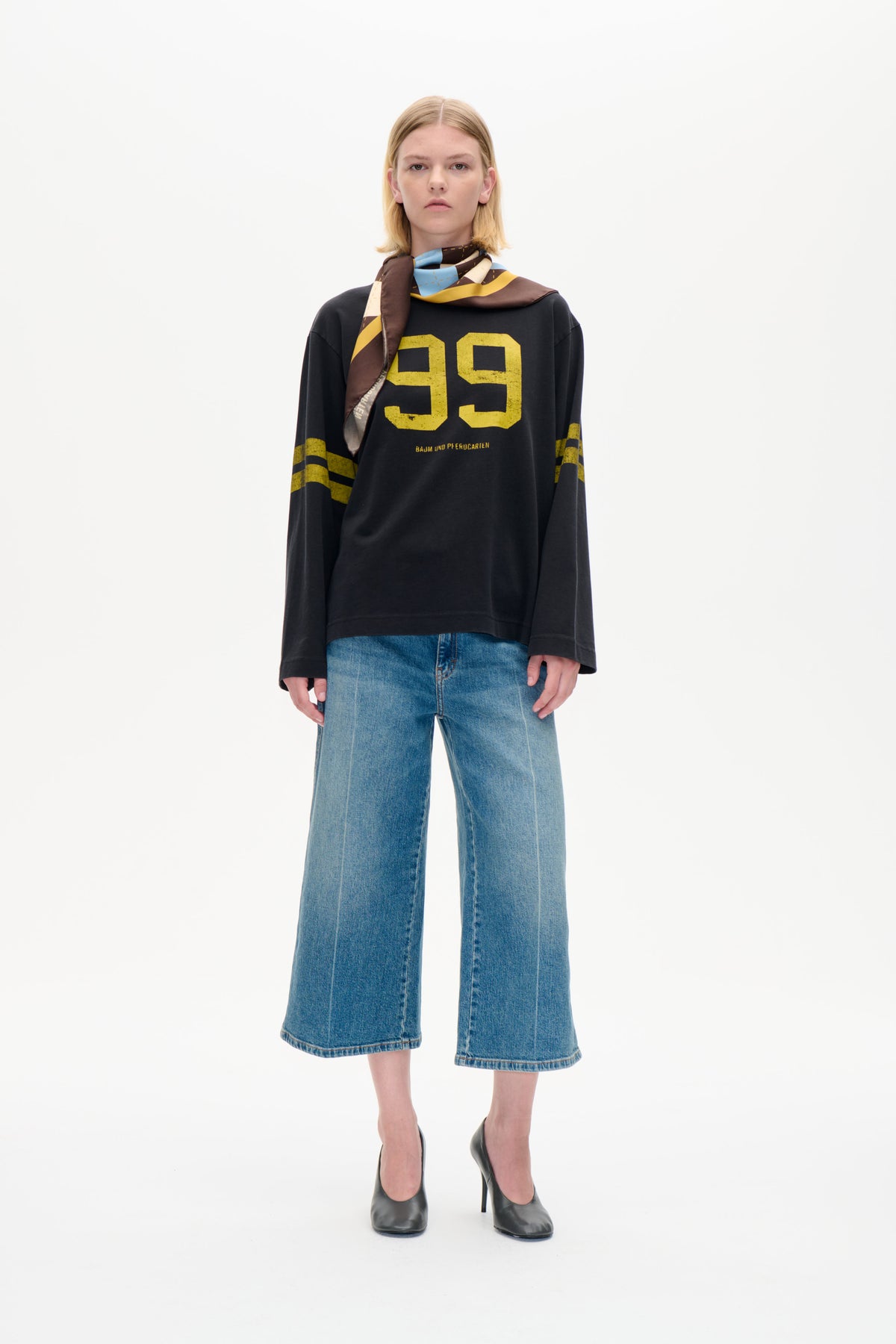 Model iført en koksgrå langærmet sweatshirt med et stort gult 99-print, blå jeans med vide ben og et flerfarvet geometrisk tørklæde.