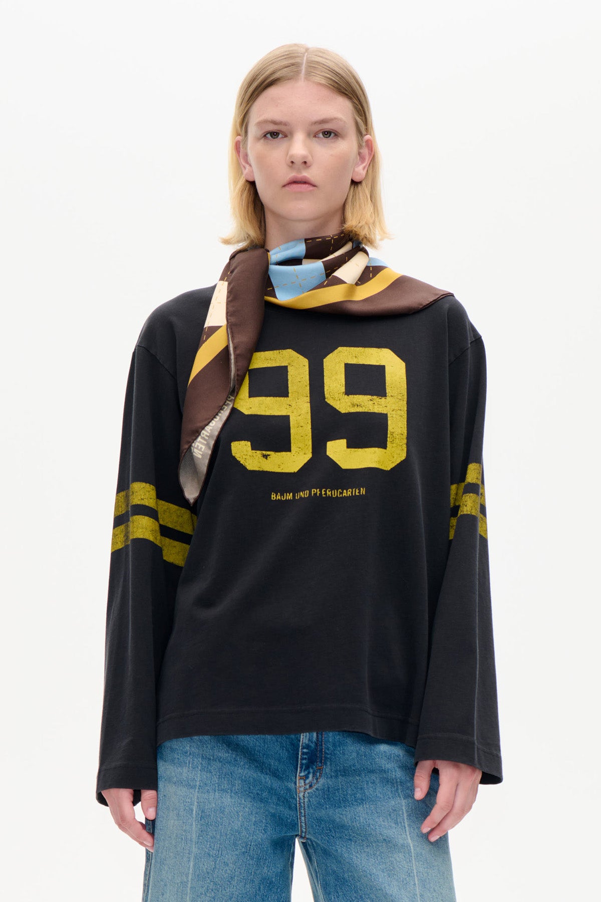 Set forfra: en model iført en sort langærmet top med gult talprint, et flerfarvet mønstret tørklæde og blå jeans.