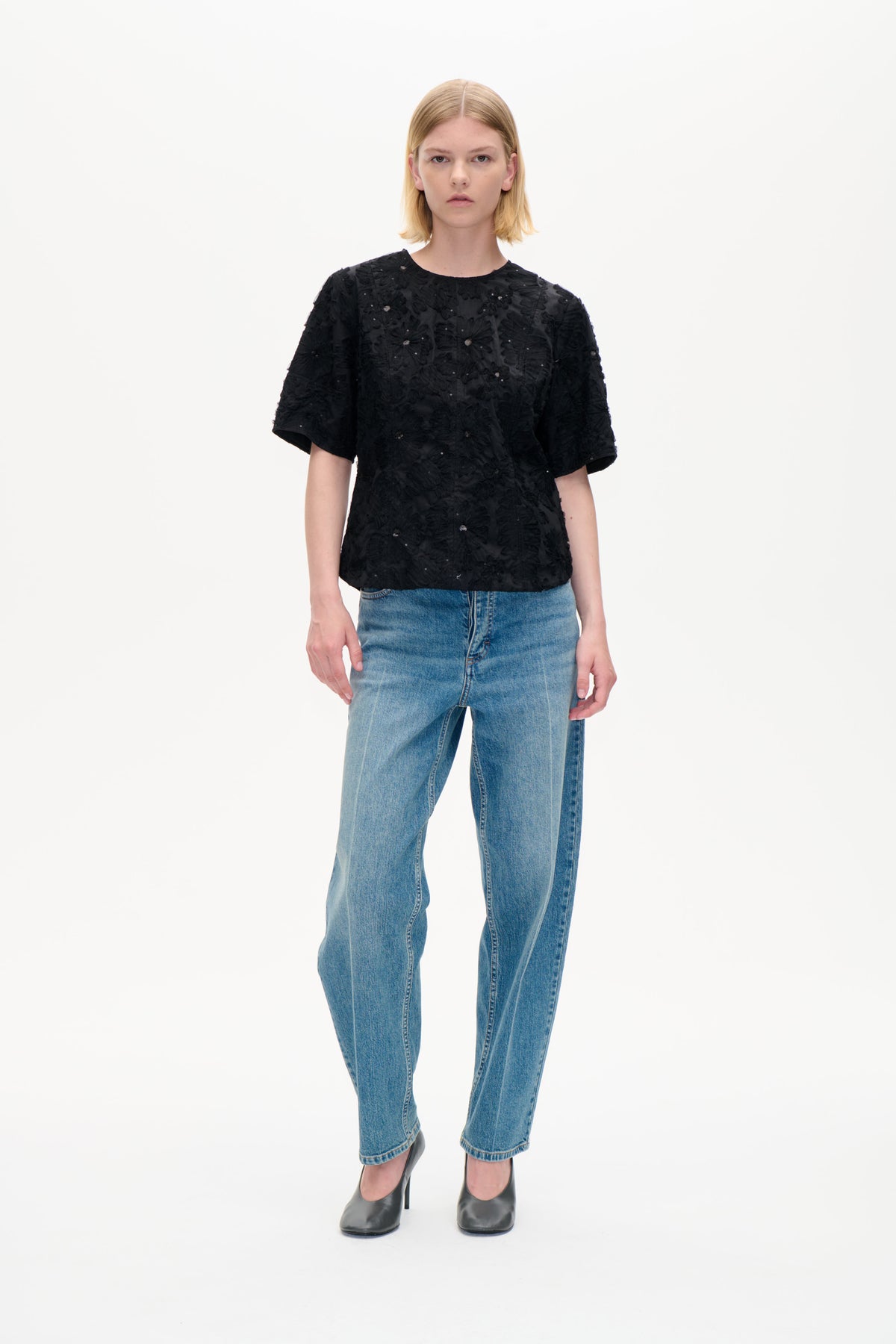 Model set forfra iført en sort kortærmet top med fremhævede blomstermotiver og små dekorative detaljer, parret med blå jeans.