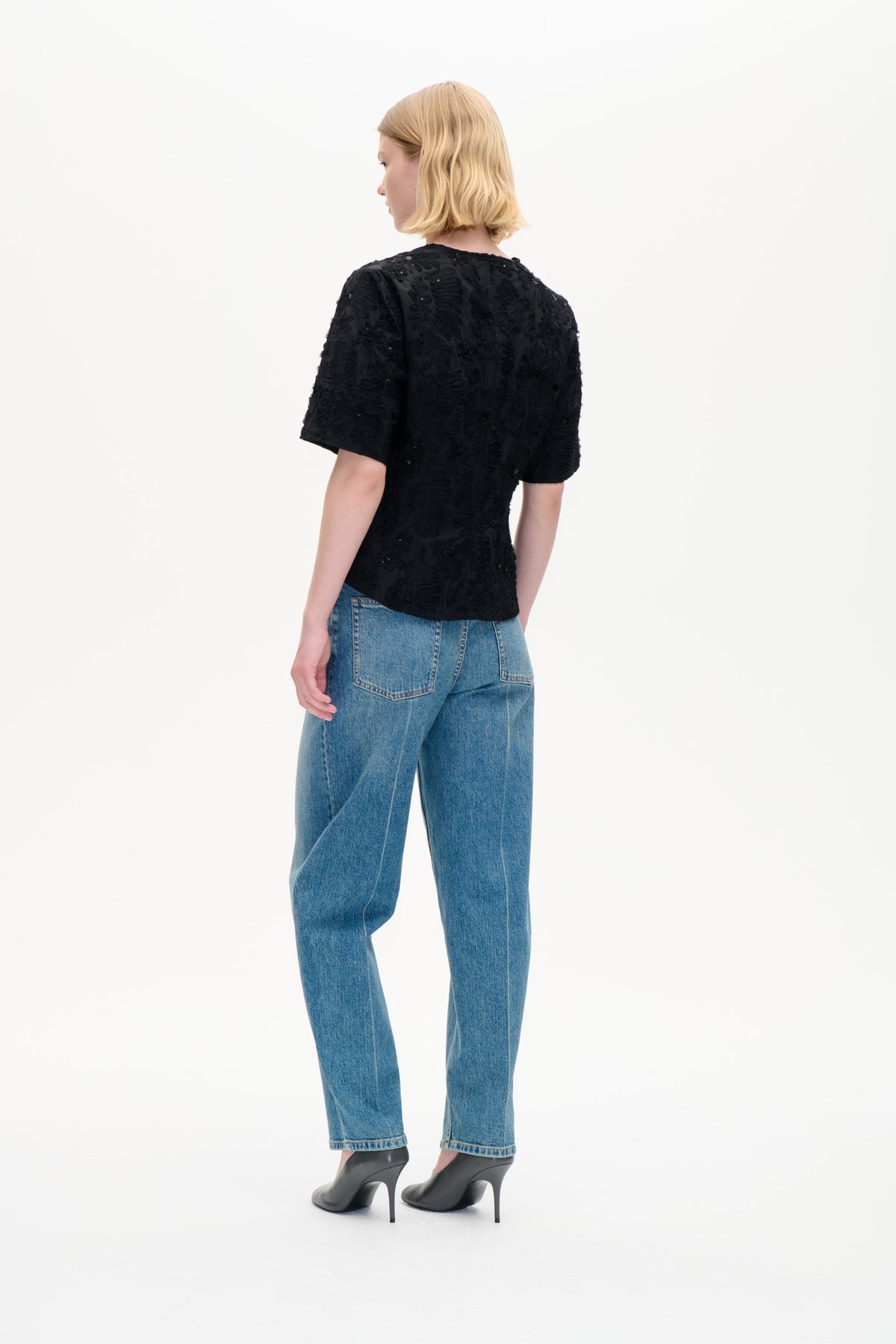 Model set bagfra iført en sort kortærmet top med tekstureret blomsterdekoration og blå jeans.