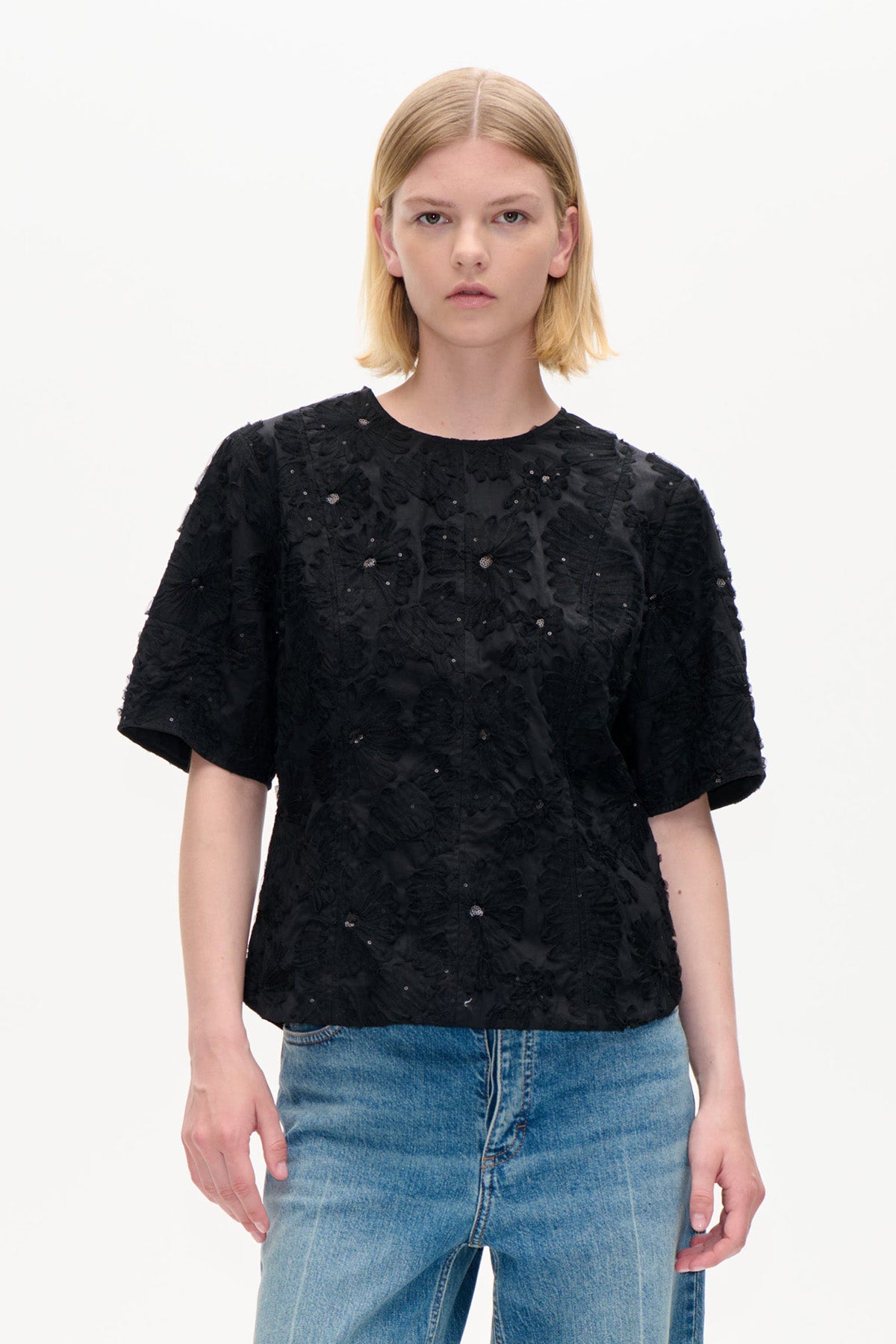Forfra: sort kortærmet top med fremhævede blomsterdetaljer og små pynteelementer, kombineret med blå jeans.