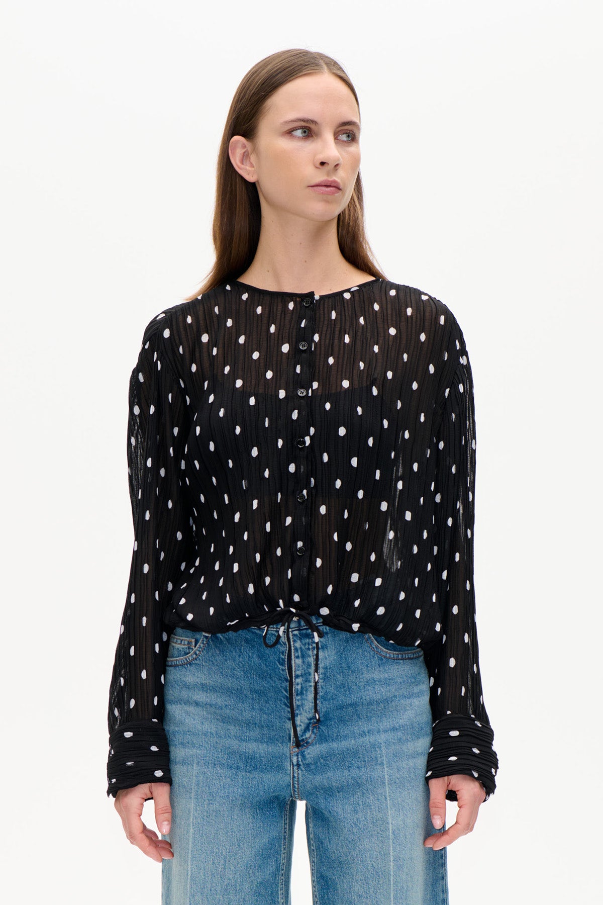 Set forfra: en sort transparent rynket bluse med hvide uregelmæssige prikker, knaplukning foran og lange ærmer, stylet med blå jeans.