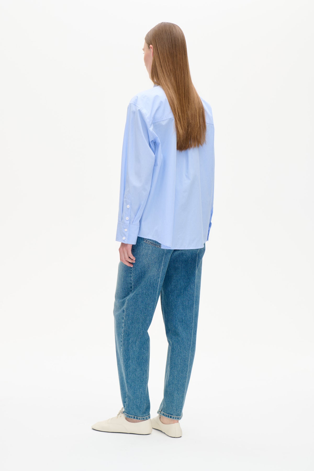 Set bagfra en model iført en lyseblå fintstribet skjorte med knapper og lange ærmer samt blå jeans.