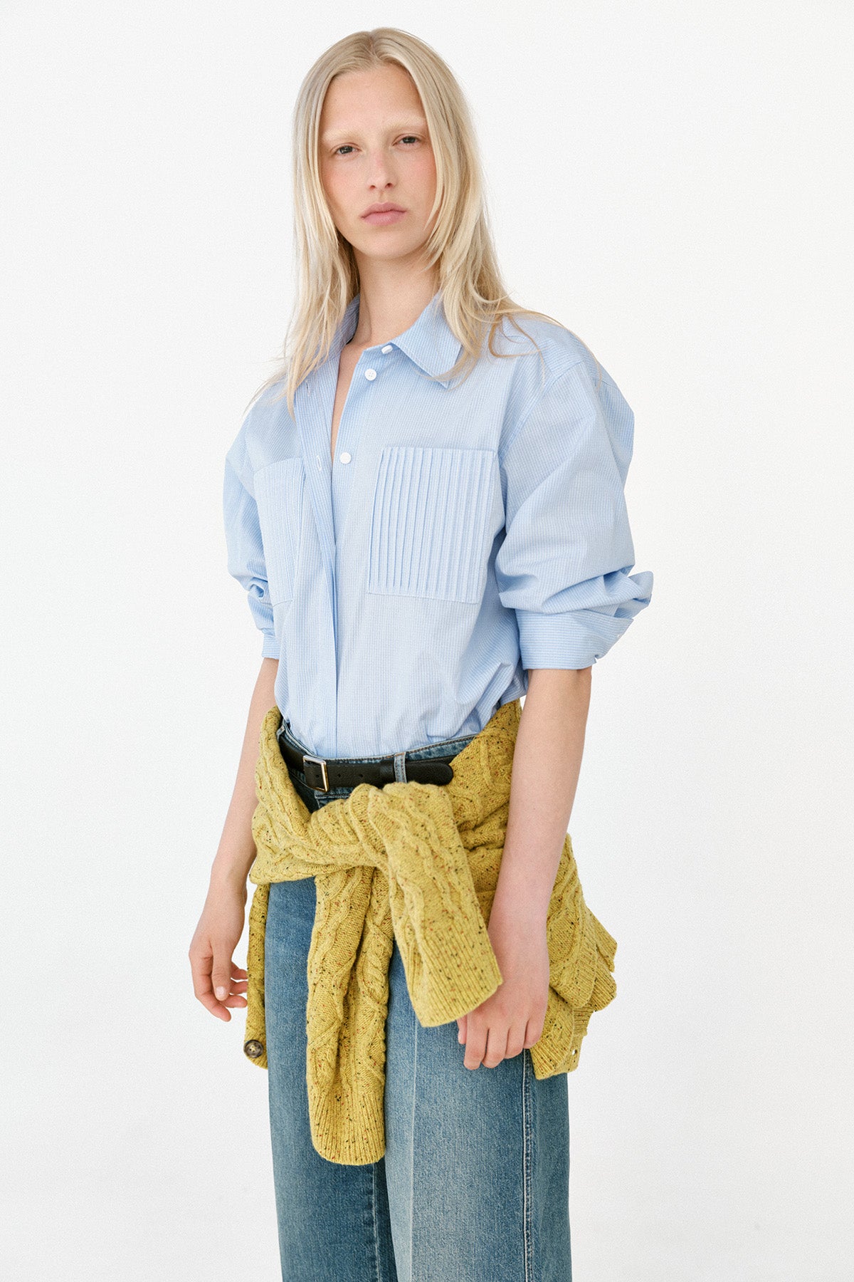 Model iført en lys blå oversized skjorte med knapper, plisseret brystlomme, vide blå jeans og en sennepsgul strik bundet i taljen.