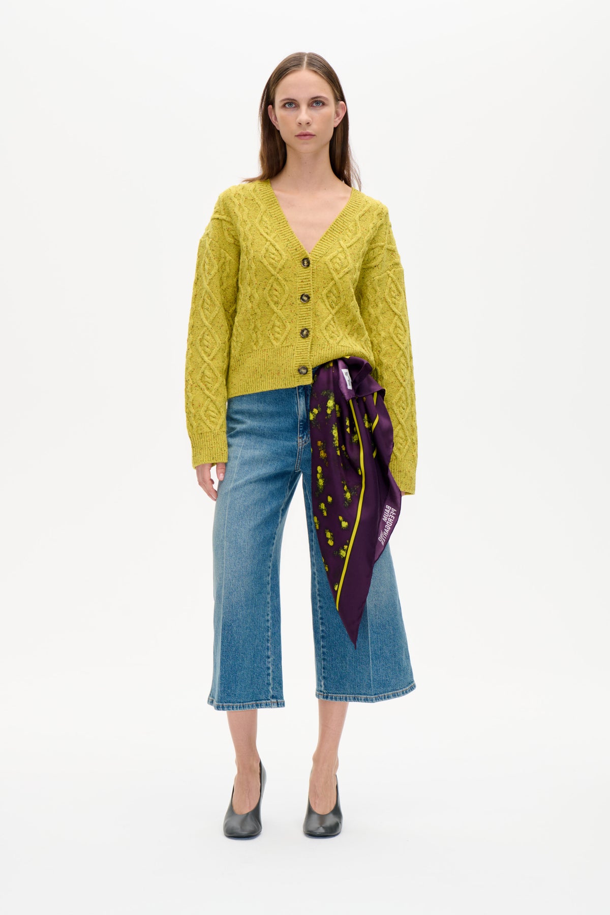Model set forfra iført en gulgrøn kabelstrikket cardigan med knapper, blå wide-leg jeans og et lilla blomstret tørklæde.