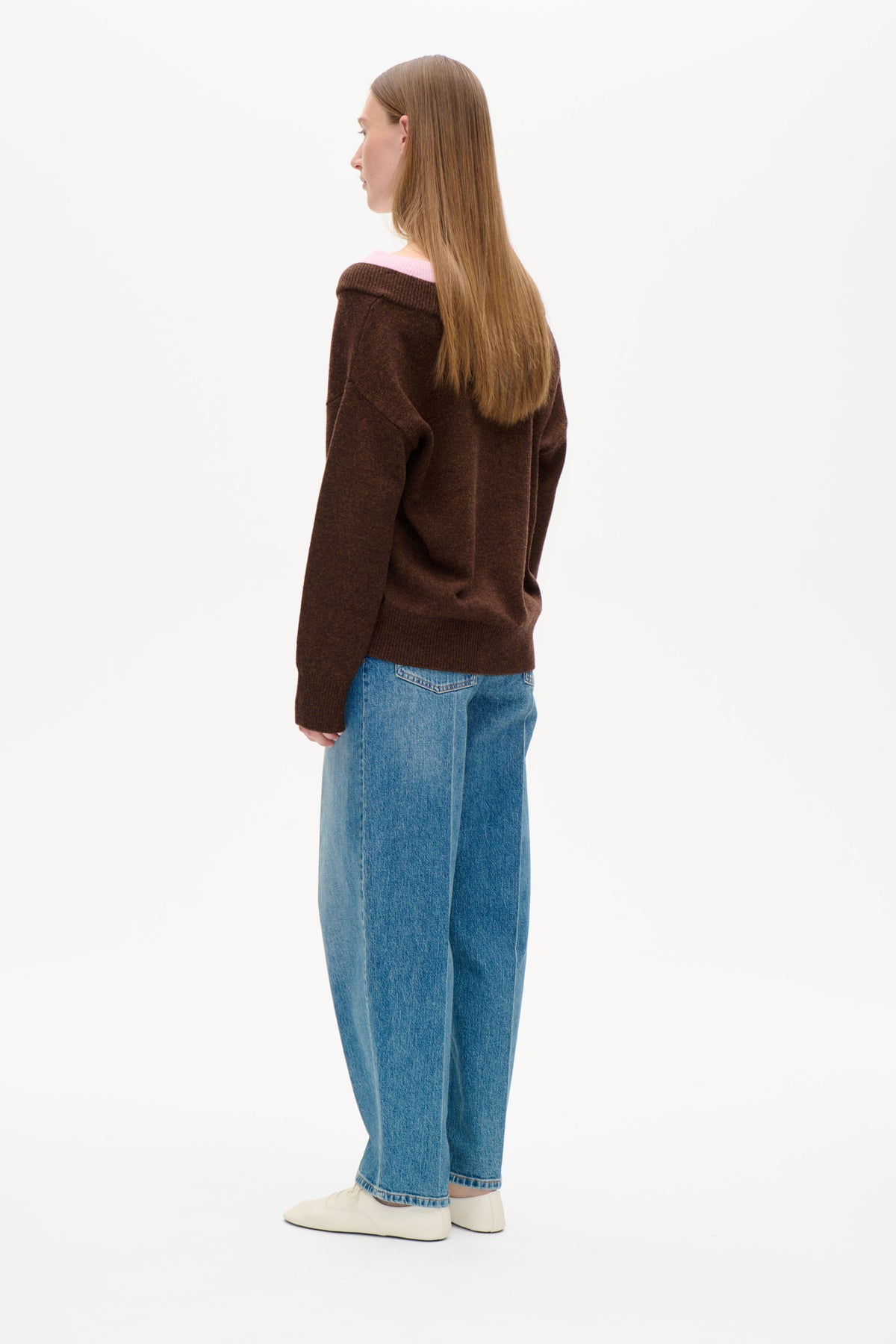 Set bagfra en model iført en mørkebrun strikket sweater med en bred, foldet halsudskæring, der viser et sart lyserødt lag.