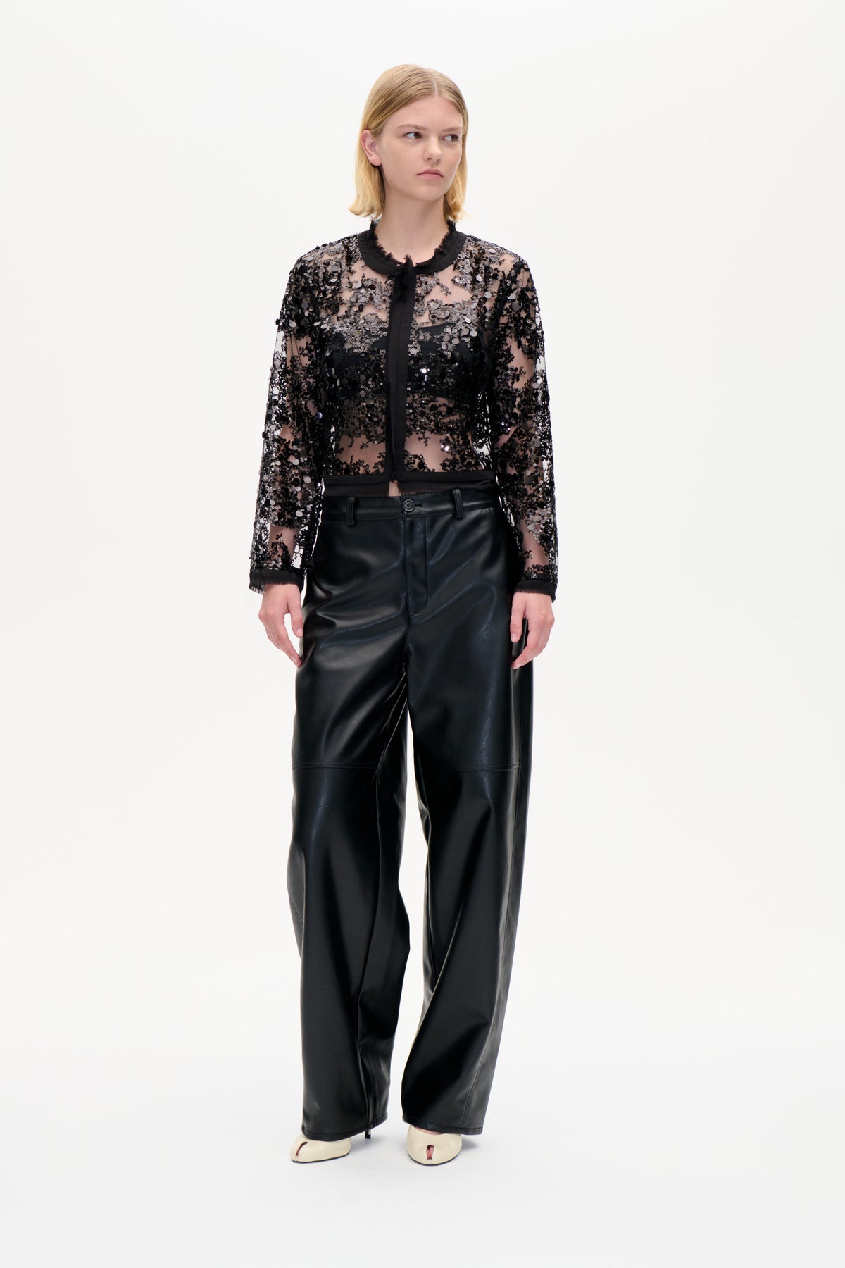 Model iført en transparent sort pailletbluse med bindebånd i kraven og sorte bukser med vide ben, vist forfra mod en hvid studiobaggrund.