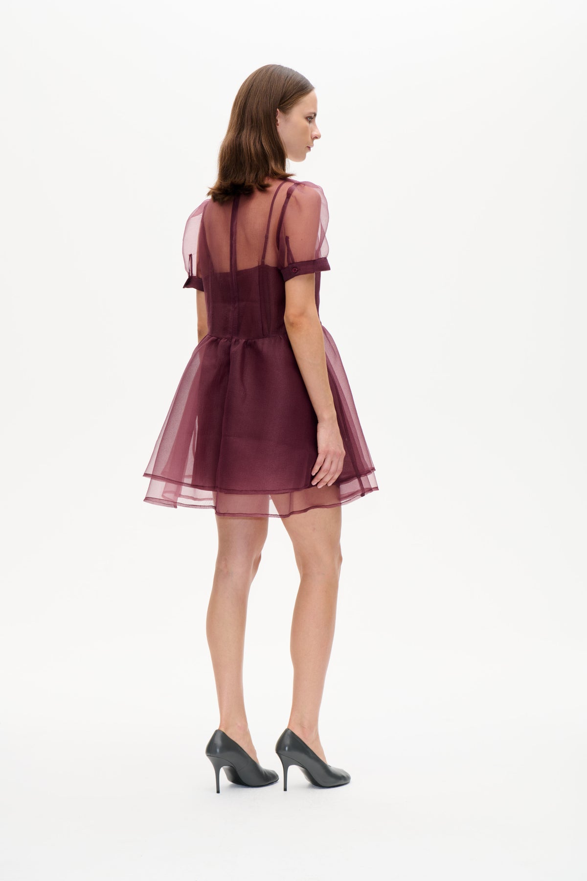 Model i en plommefarvet minikjole med let gennemsigtige mauve pufærmer og en lagdelt, transparent nederdel, set bagfra i trekvart vinkel.