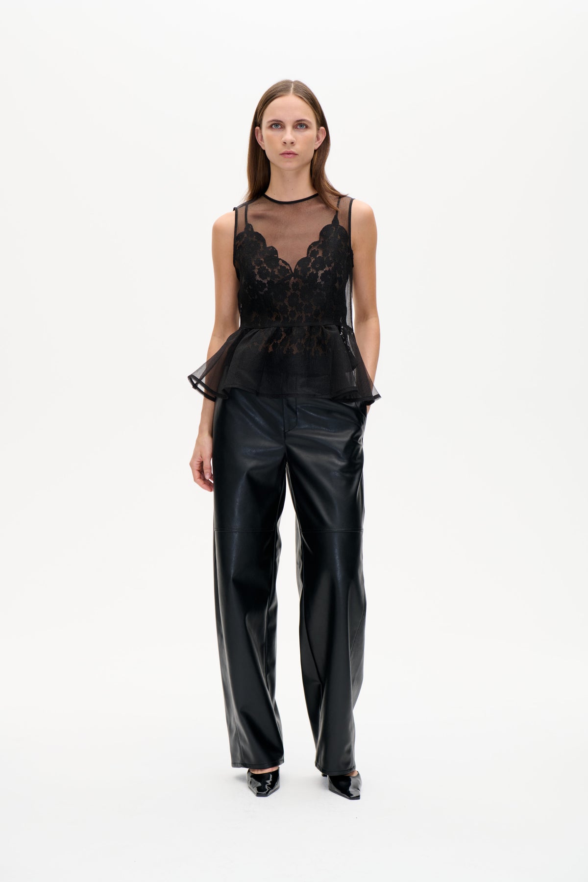 Model iført en sort, transparent ærmeløs top med blomstrede blondepaneler og et peplum med vidde, stylet med blanke bukser med vide ben.